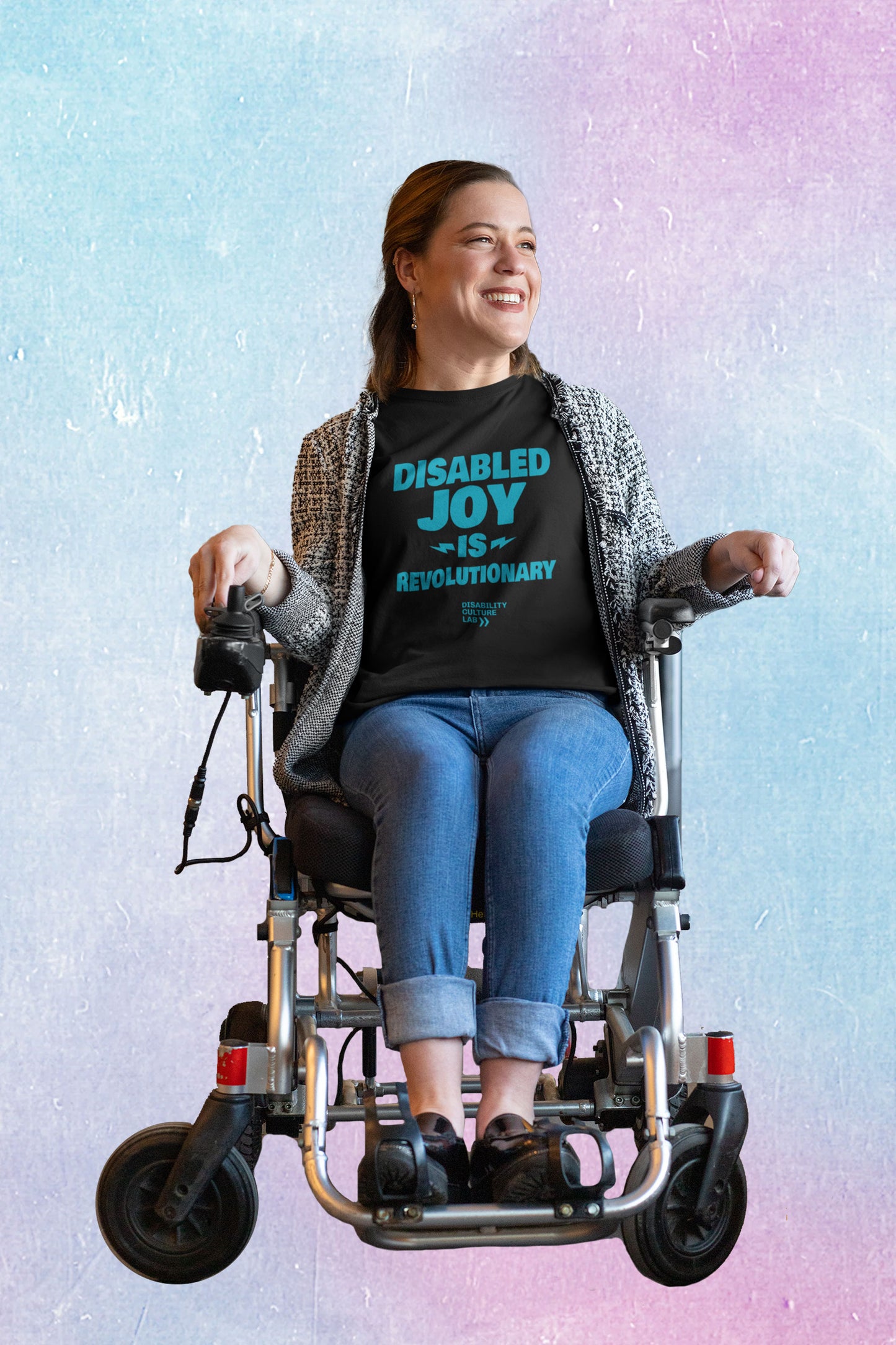 A person with brown hair sits in a power chair, looking to the left with a bright smile. They are wearing a gray sweater, blue jeans, and a black t-shirt with the turquoise words "Disabled Joy Is Revolutionary" and the turquoise Disability Culture Lab logo.