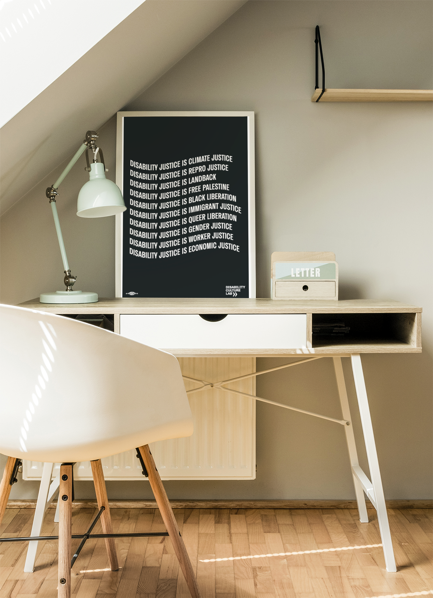 A black poster with the words: "Disability Justice Is... Climate Justice, Repro Justice, Landback, Free Palestine, Black Liberation, Immigrant Justice, Queer Liberation, Gender Justice, Worker Justice, Economic Justice" in a white frame on a white and beige desk, beside a white table lamp and chair.