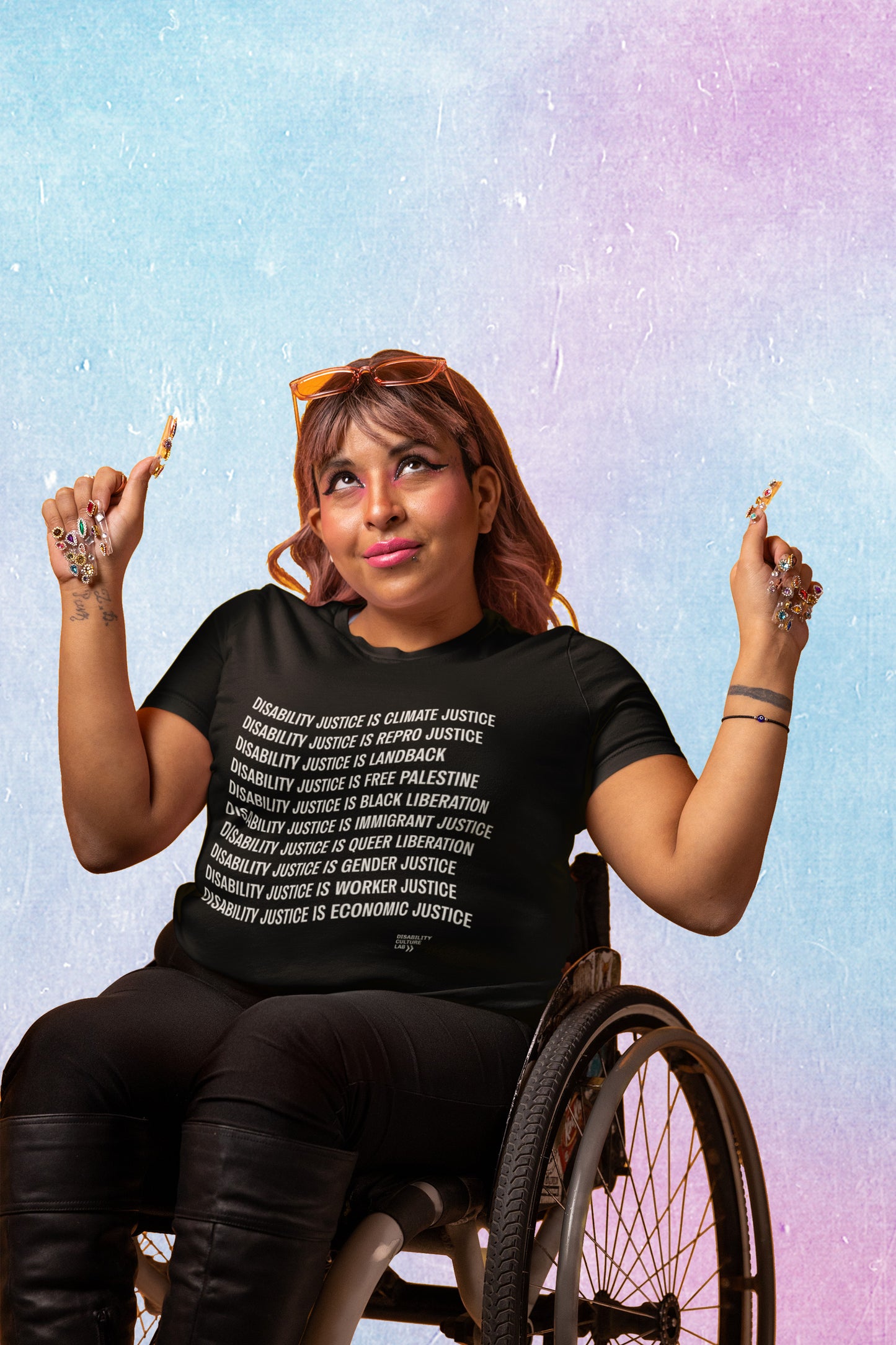 A brown-skinned person with auburn hair, long nails, and amber-tinted sunglasses sits in a wheelchair while wearing a black Disability Culture Lab shirt, Union Printed and Made in the USA, deisgned by Jennifer White-Johnson: Text reads: "Disability Justice Is... Climate Justice, Repro Justice, Landback, Free Palestine, Black Liberation, Immigrant Justice, Queer Liberation, Gender Justice, Worker Justice, Economic Justice."