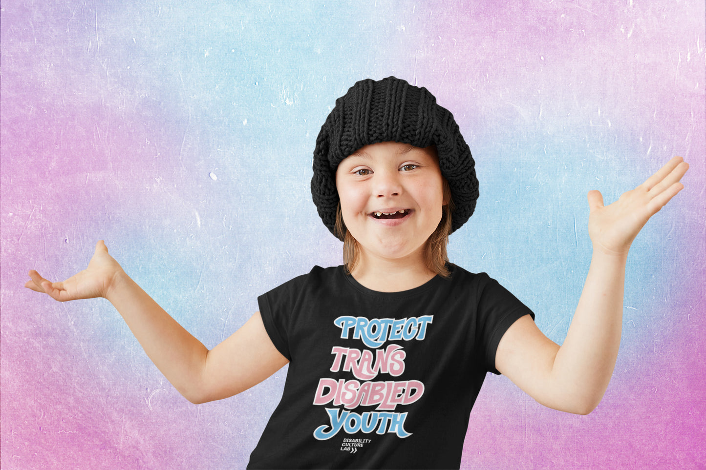A very cute smiling white child in a black beanie and black t-shirt that reads “Protect Trans Disabled Youth” in hand drawn letters in the trans pride flag colors (light pink, sky blue, and white).