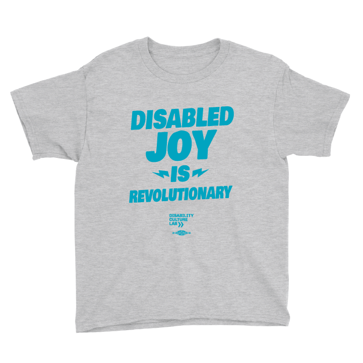 A very cute white child is blowing bubbles while wearing blue jeans shorts and a gray shirt with the turquoise words "Disabled Joy Is Revolutionary" and the turquoise Disability Culture Lab logo.