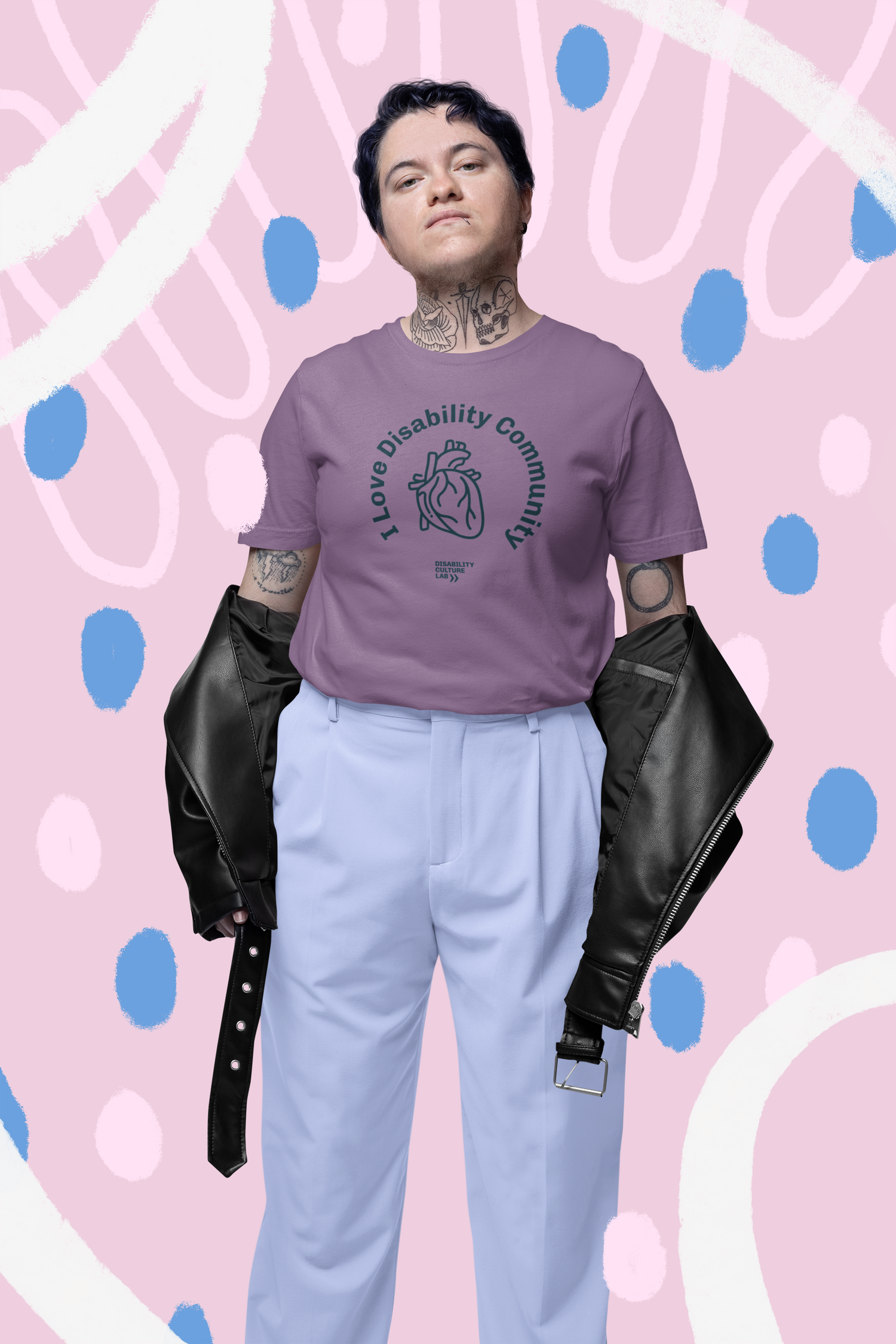 Person with tattoos wearing a lavender t-shirt with "I love Disability Community" text, light blue pants, and a black jacket draped over their arms. The background is pink with white and blue abstract shapes.