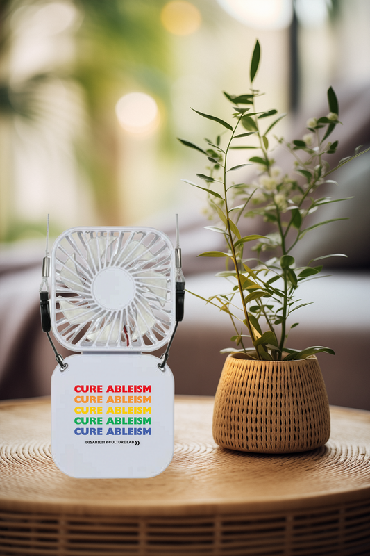 A small white portable fan that reads "Cure Ableism" five times in red, orange, yellow, green, and blue, and the white Disability Culture Lab logo sitting on a wooden table next to a plant.