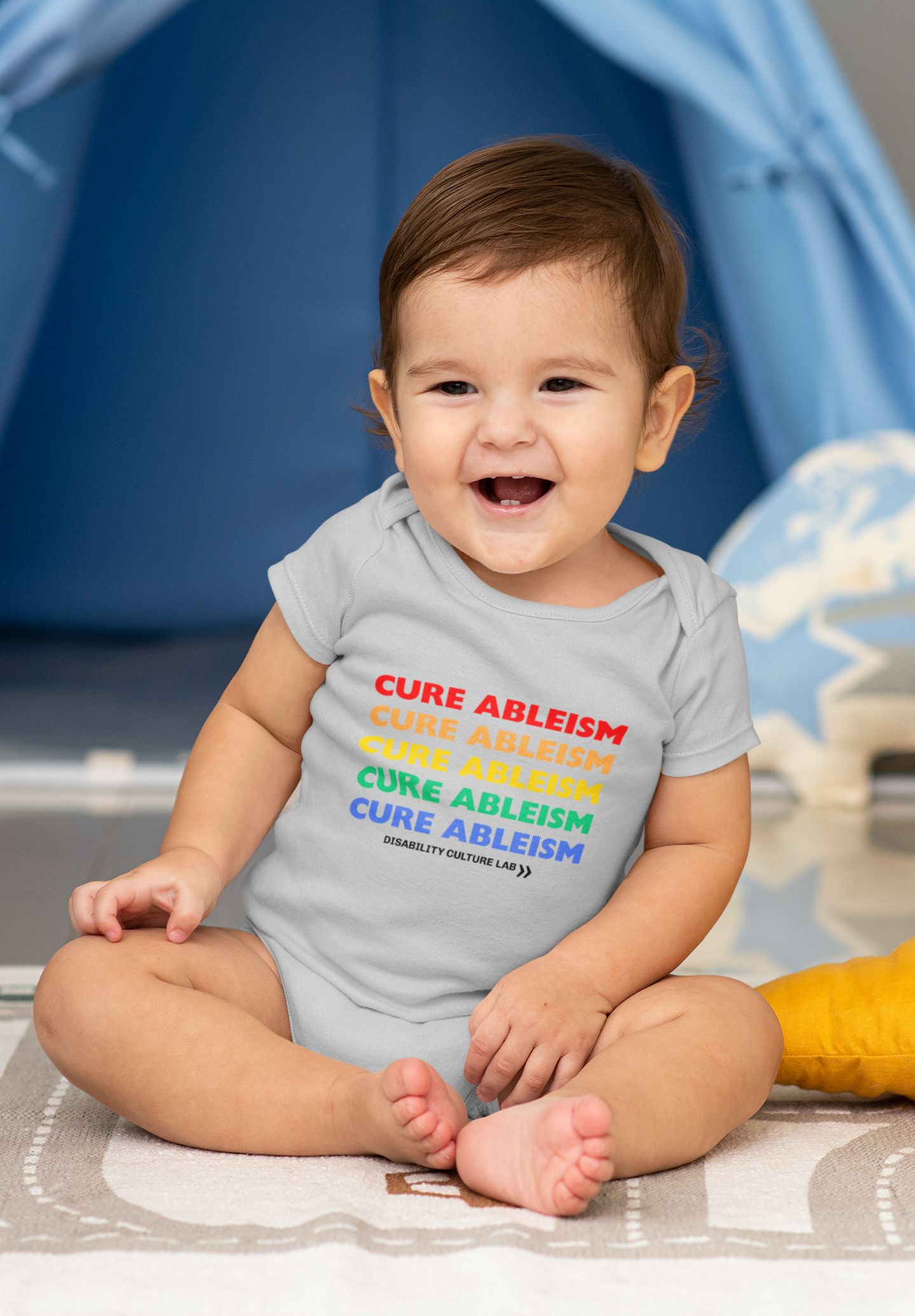 A very cute white baby sits while wearing a gray onesie that reads "Cure Ableism" five times in red, orange, yellow, green, and blue, and the white Disability Culture Lab logo. 