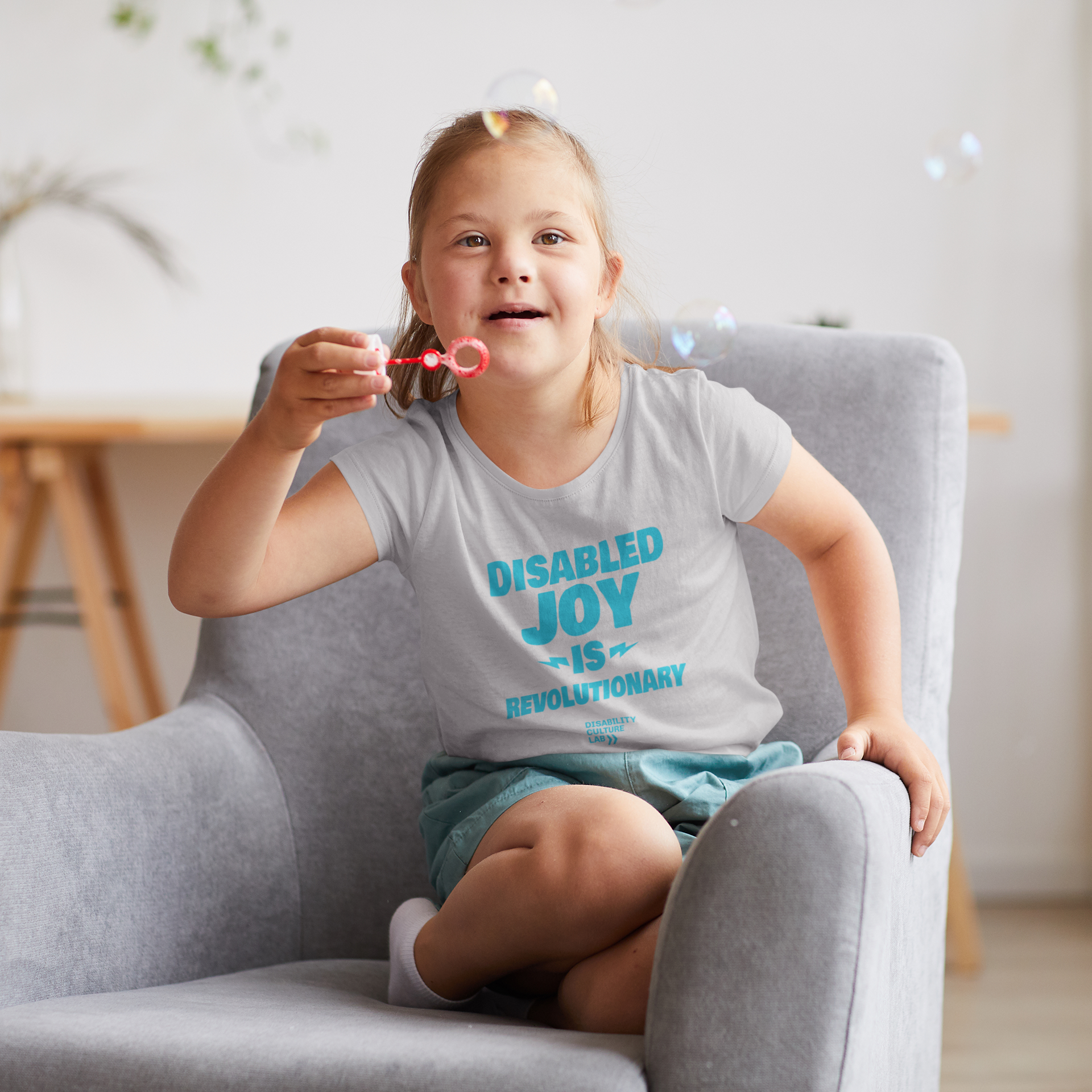 A very cute white child is blowing bubbles while wearing a gray shirt with the turquoise words "Disabled Joy Is Revolutionary" and the turquoise Disability Culture Lab logo.