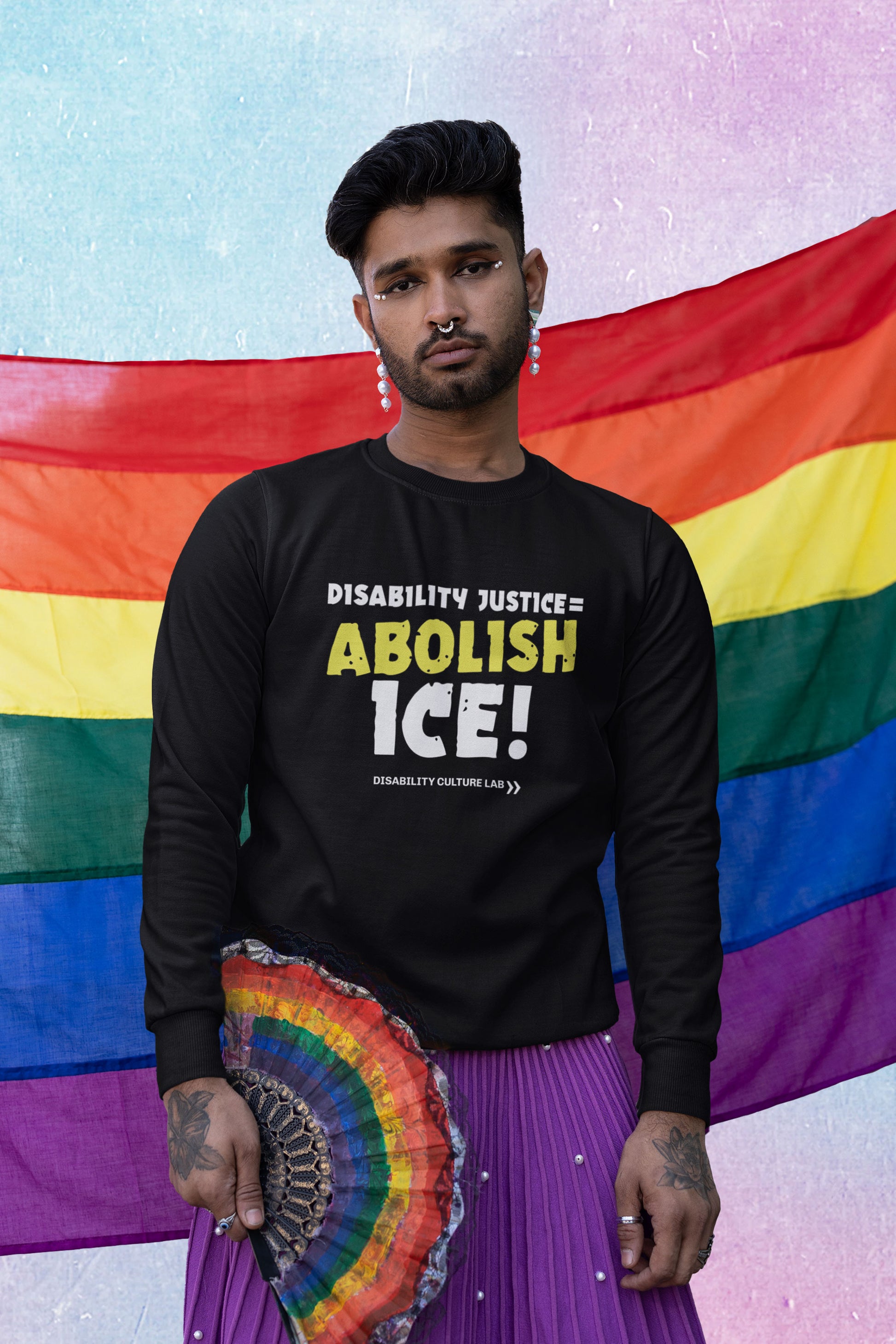 A brown-skinned person with beautiful white eye makeup, pearl-drop earrings, and a septum ring stands facing the camera. They are holding a rainbow pride fan, and are wearing a purple skirt and a black crewneck sweater that reads "Disability Justice = Abolish ICE!" in mostly white text, with "Abolish" in bright yellow and slightly tilted. Below is the white Disability Culture Lab logo.
