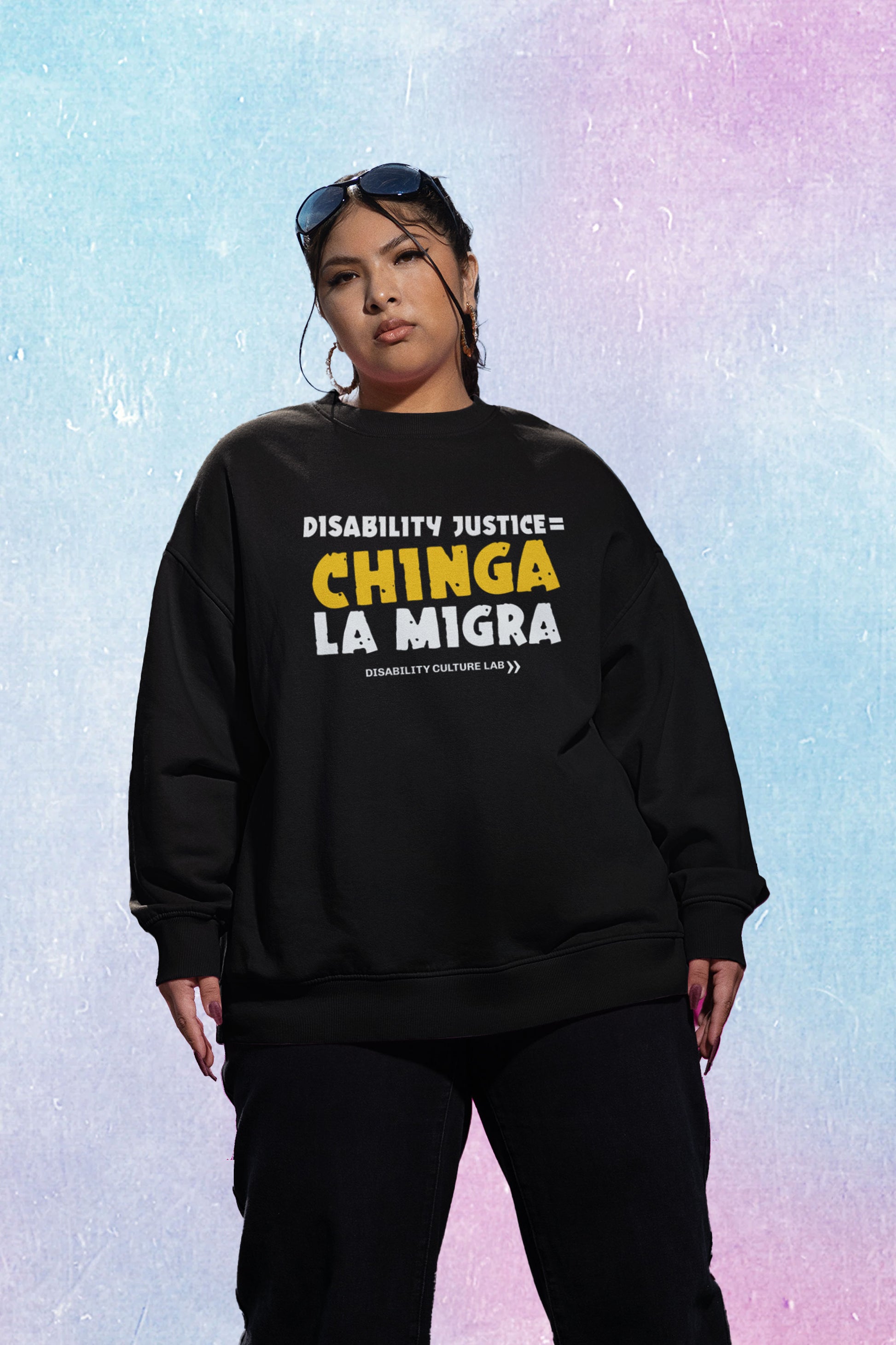 A brown-skinned person stands facing toward the camera. They are wearing sunglasses on top of their head, hoop earrings, black pants, and a black crewneck sweater that reads "Disability Justice = Chinga La Migra" in mostly white text, with "Chinga" in bright yellow and slightly tilted. Below is the white Disability Culture Lab logo.