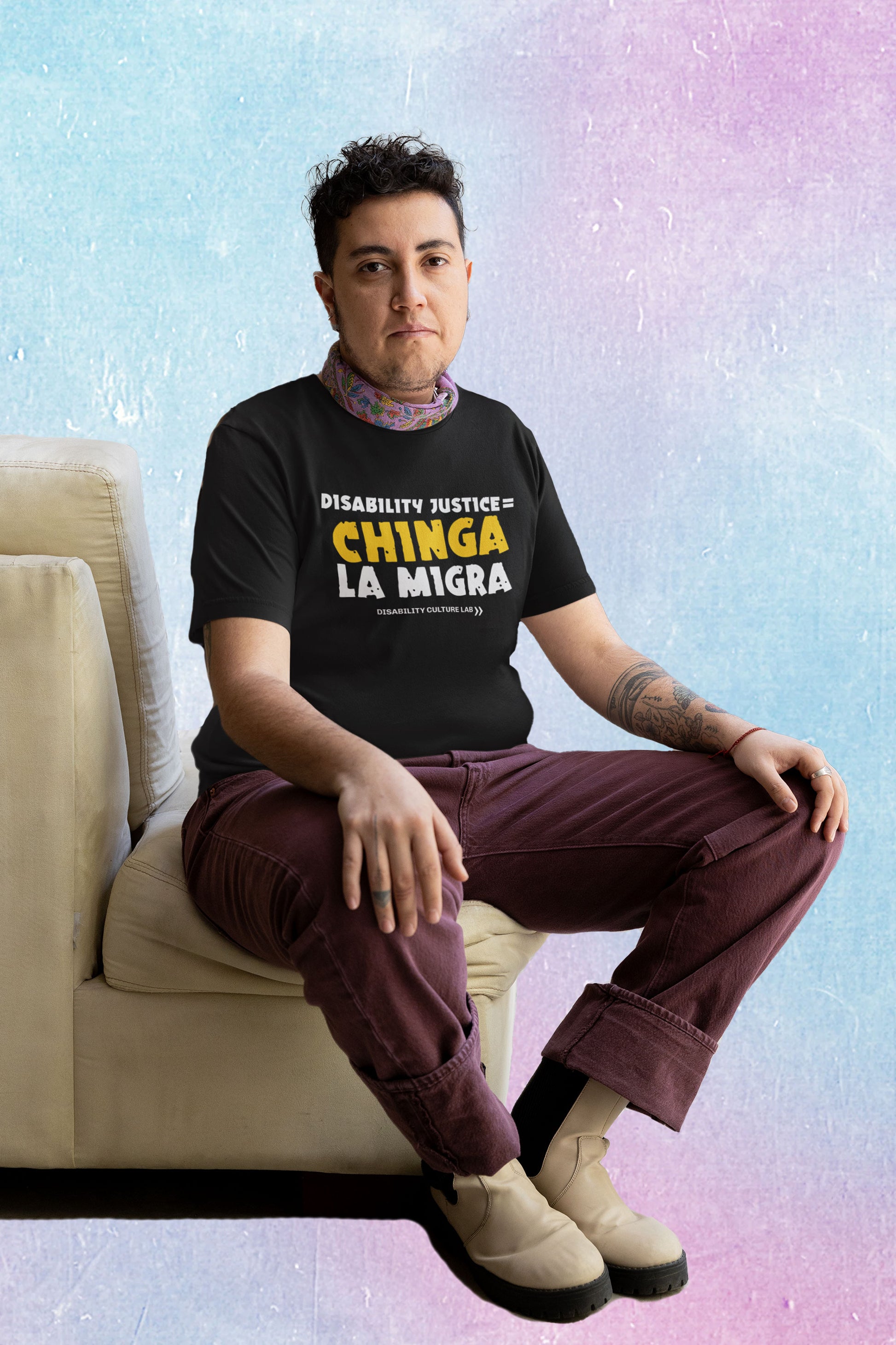A person with black, cropped, curly hair sits on a tan chair cushion, looking at the camera. They are wearing brown pants, white boots, and a black t-shirt that reads "Disability Justice = Chinga La Migra" in mostly white text, with "Chinga" in bright yellow and slightly tilted. Below is the white Disability Culture Lab logo.