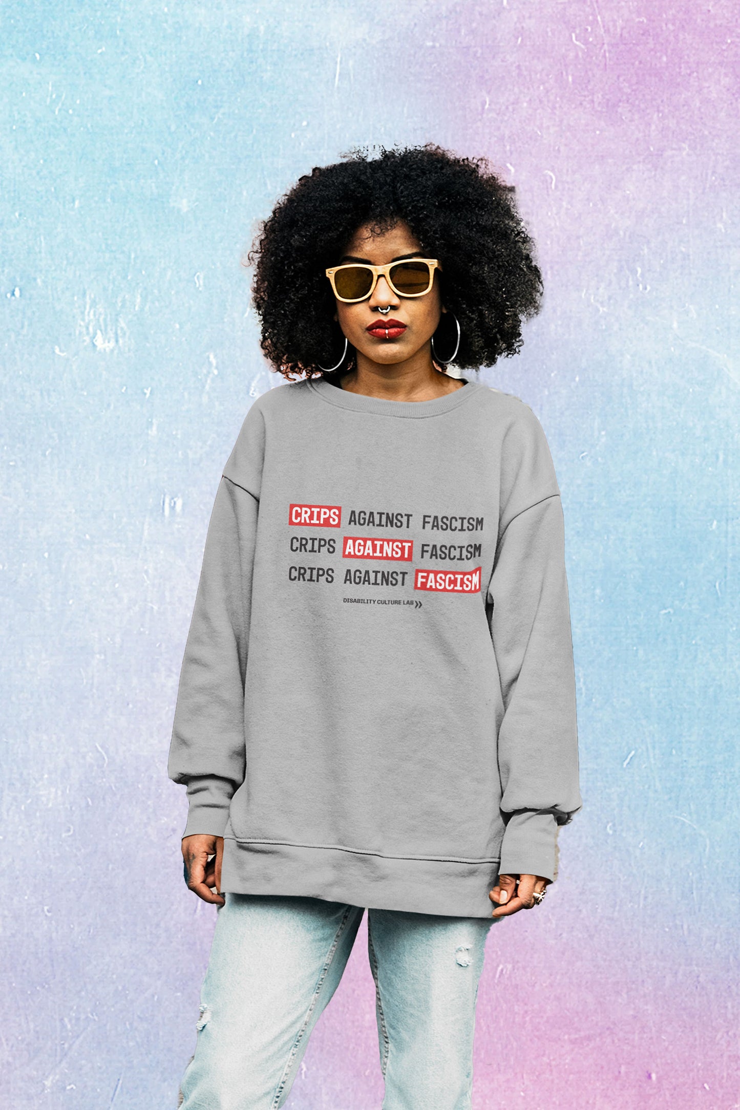 A Black person with big, black, curly hair, tan-tined sunglasses, a septum ring, lip ring, red lipstick, and silver hoop earrings faces the camera. They are wearing a gray crewneck sweater that reads "Crips Against Fascism" three times, with "Crips" in the first line, "Against" in the second line, and "Fascism" in the third line all in white text with bright red background, so it also reads "Crips Against Fascism" diagonally. Below is the black Disability Culture Lab logo.