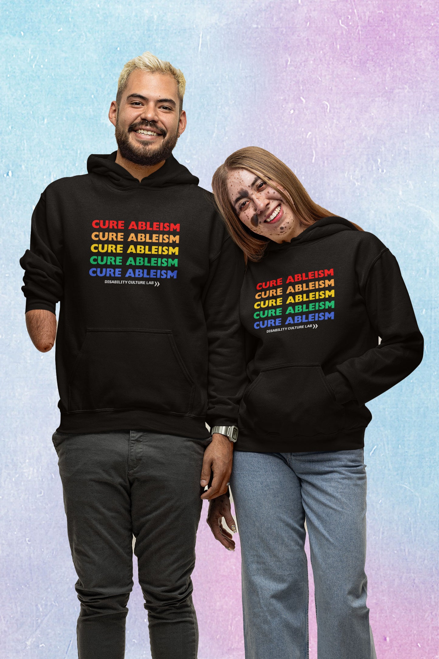Two people—one with limb difference and one with a facial birthmark—stand smiling at the camera, wearing black pullover hoodies that read "Cure Ableism" five times in red, orange, yellow, green, and blue, and the white Disability Culture Lab logo. 