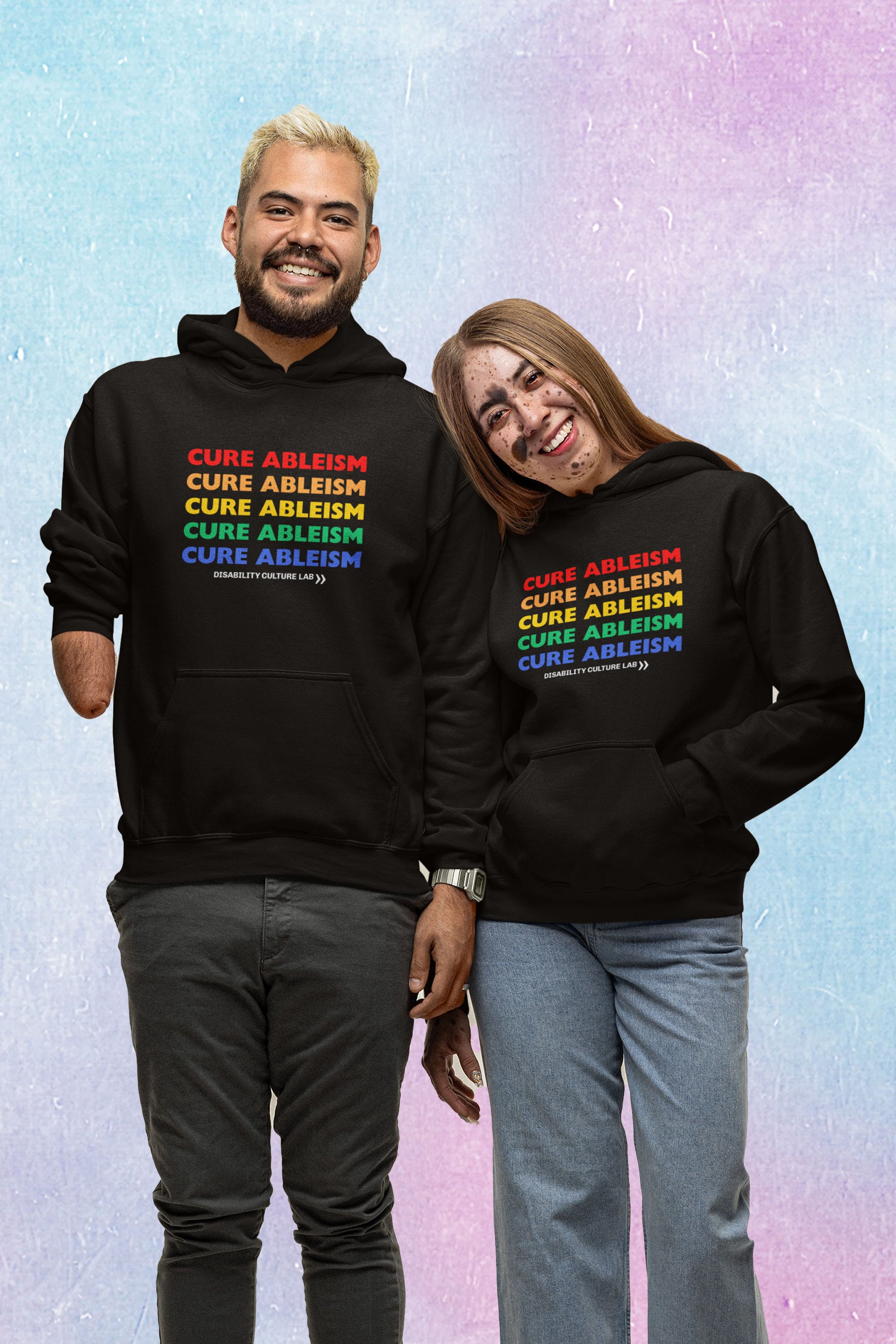 Two people—one with limb difference and one with a facial birthmark—stand smiling at the camera, wearing black pullover hoodies that read "Cure Ableism" five times in red, orange, yellow, green, and blue, and the white Disability Culture Lab logo. 