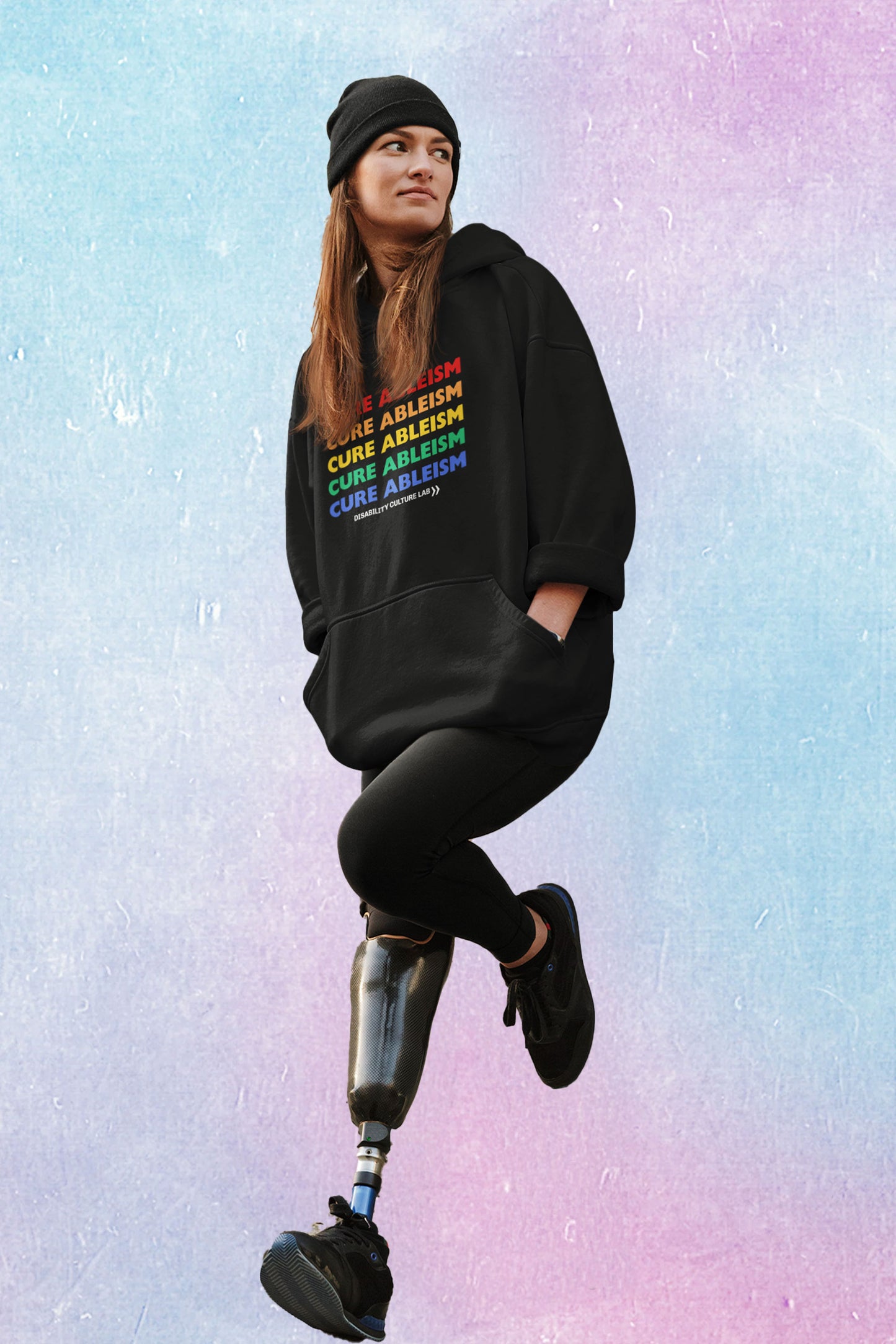 A person with long brown hair and a black prosthetic leg poses, looking off to the left. They are wearing a black beanie, black leggings, black shoes, and a black pullover hoodie that reads "Cure Ableism" five times in red, orange, yellow, green, and blue, and the white Disability Culture Lab logo. 