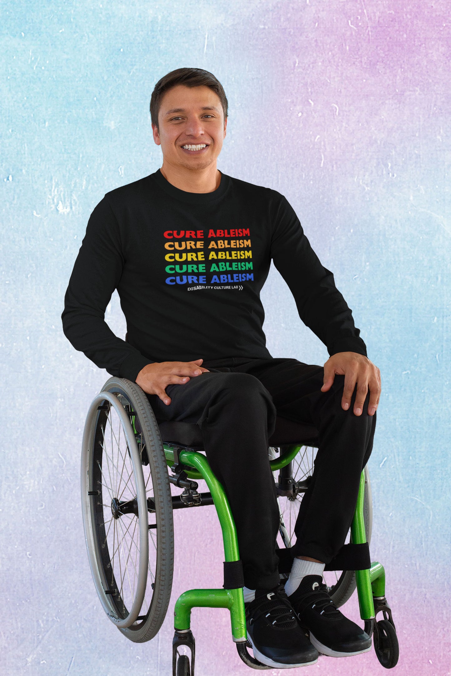 A person with short cropped dark brown hair smiles brightly into the camera, seated in a wheelchair with bright green accents. They are wearing black pants, black shoes, and a black long-sleeved shirt that reads "Cure Ableism" five times in red, orange, yellow, green, and blue, and the white Disability Culture Lab logo. 