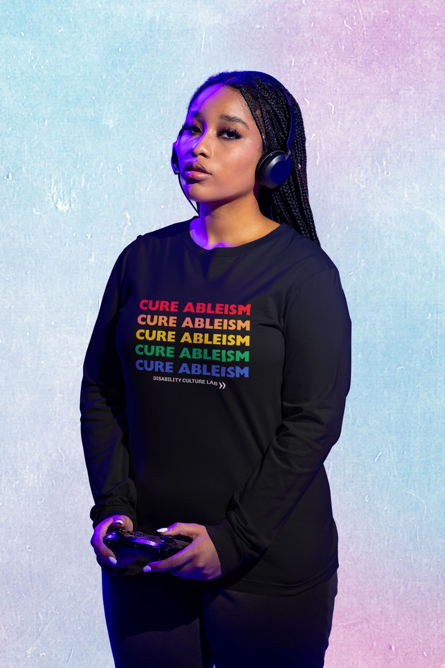 A brown-skinned Black person with long black braids looks into the camera. They are holding a gaming controller and wearing black headphones, black pants, and a black long-sleeved shirt that reads "Cure Ableism" five times in red, orange, yellow, green, and blue, and the white Disability Culture Lab logo. 