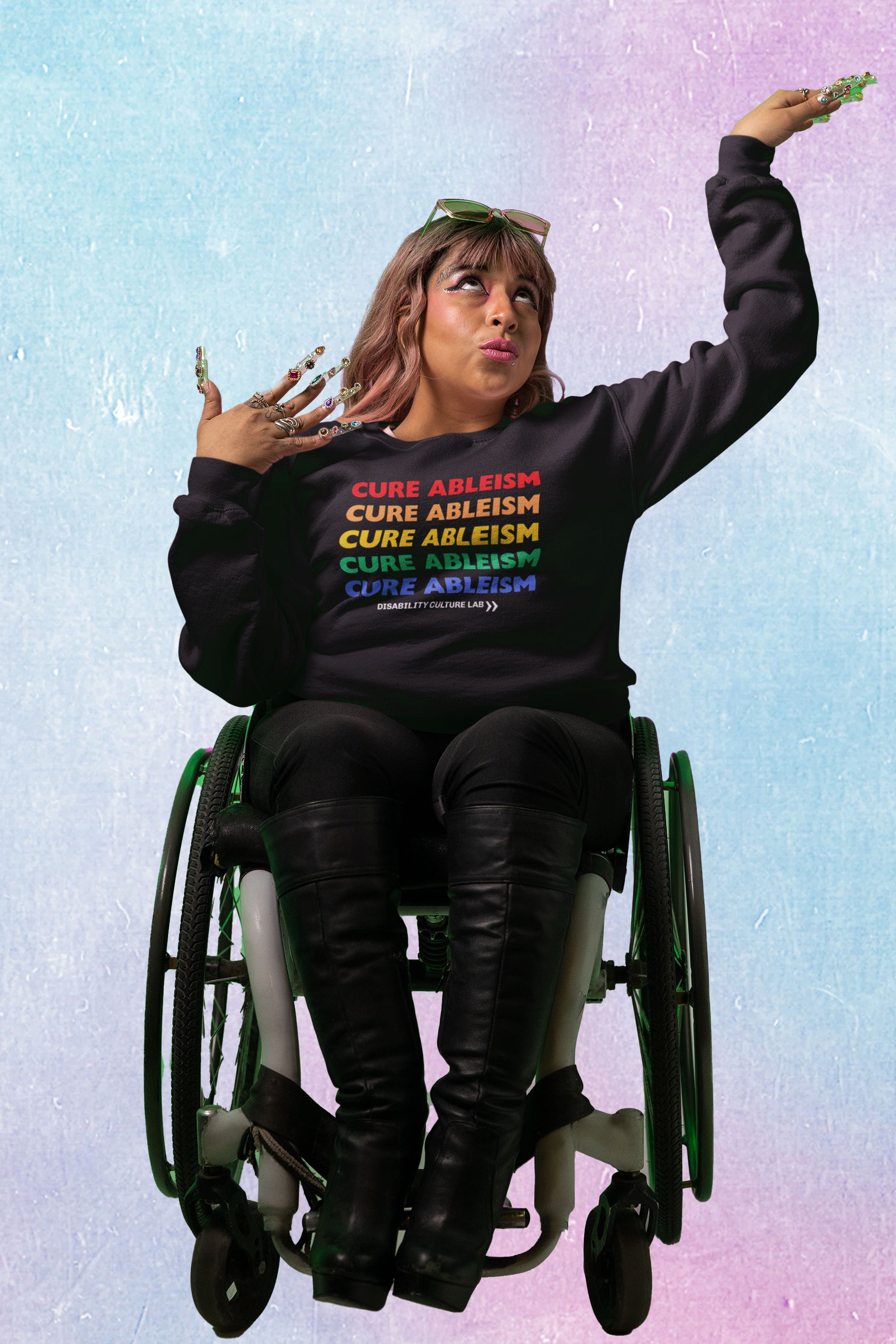 A person with brown hair and bangs, long nails, pink tinted sunglasses, and winged eyeliner poses with their arm raised and looking toward the sky, seated in a wheelchair. They are wearing black knee-high boots and black pants, and a black crewneck sweater that reads "Cure Ableism" five times in red, orange, yellow, green, and blue, and the white Disability Culture Lab logo. 