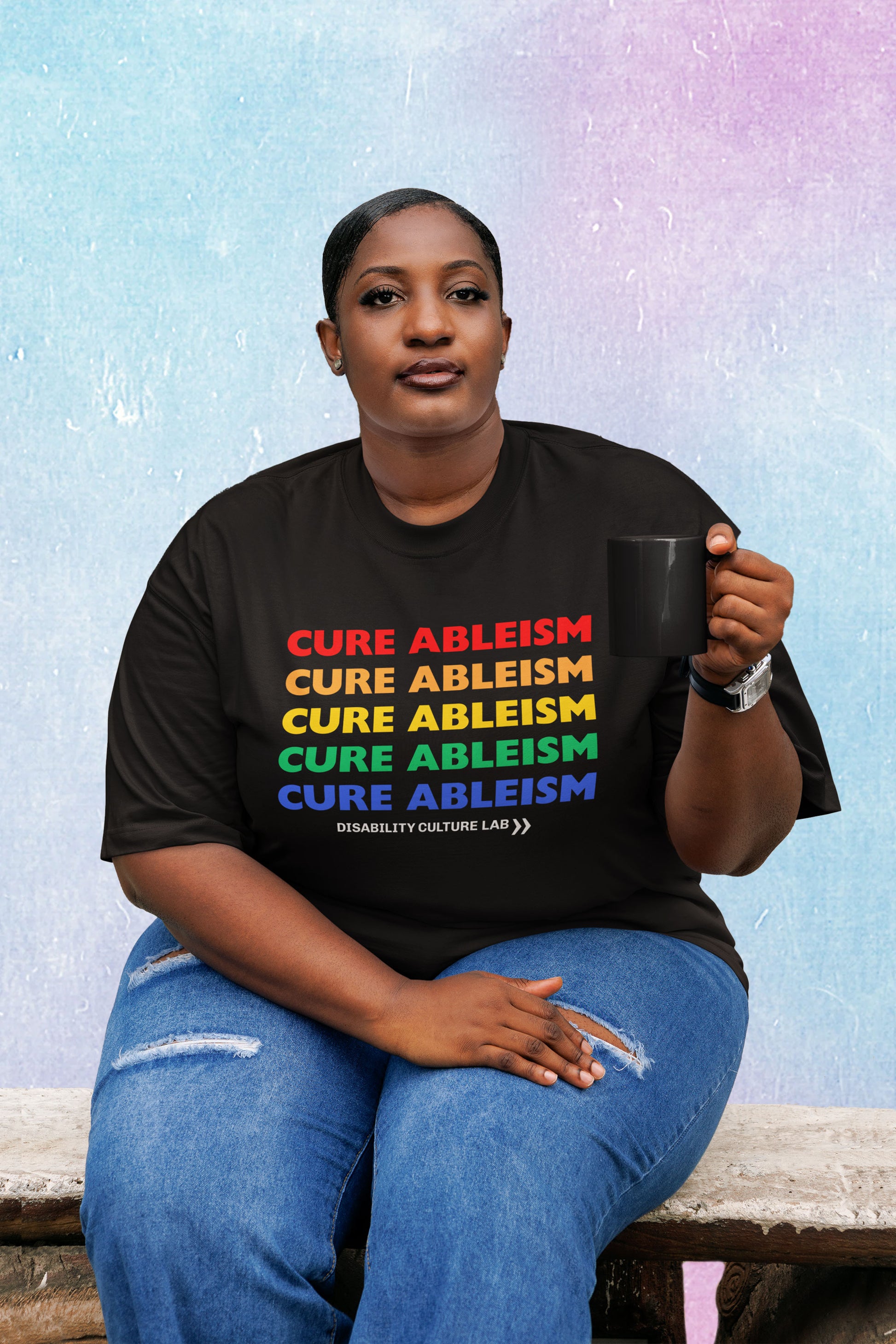 A Black person sits on a wood bench with a mug in hand, wearing ripped blue jeans and a black t-shirt that reads "Cure Ableism" five times in red, orange, yellow, green, and blue, and the white Disability Culture Lab logo. 