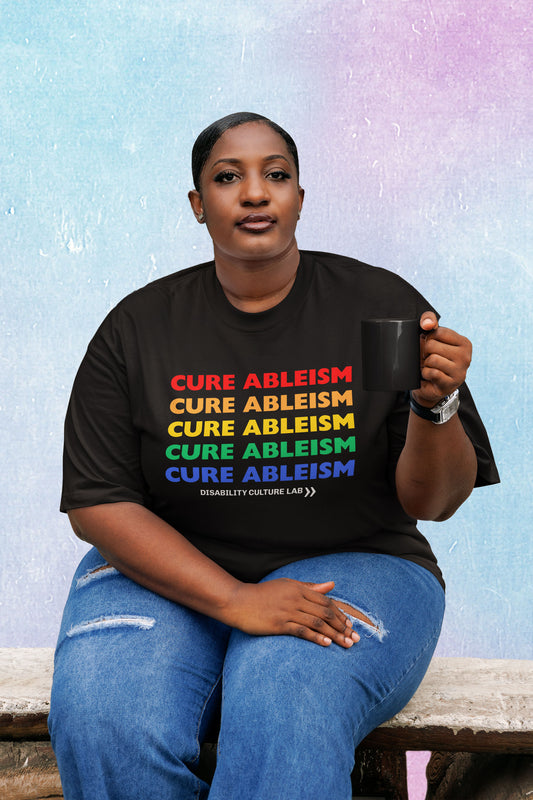 A Black person sits on a wood bench with a mug in hand, wearing ripped blue jeans and a black t-shirt that reads "Cure Ableism" five times in red, orange, yellow, green, and blue, and the white Disability Culture Lab logo. 