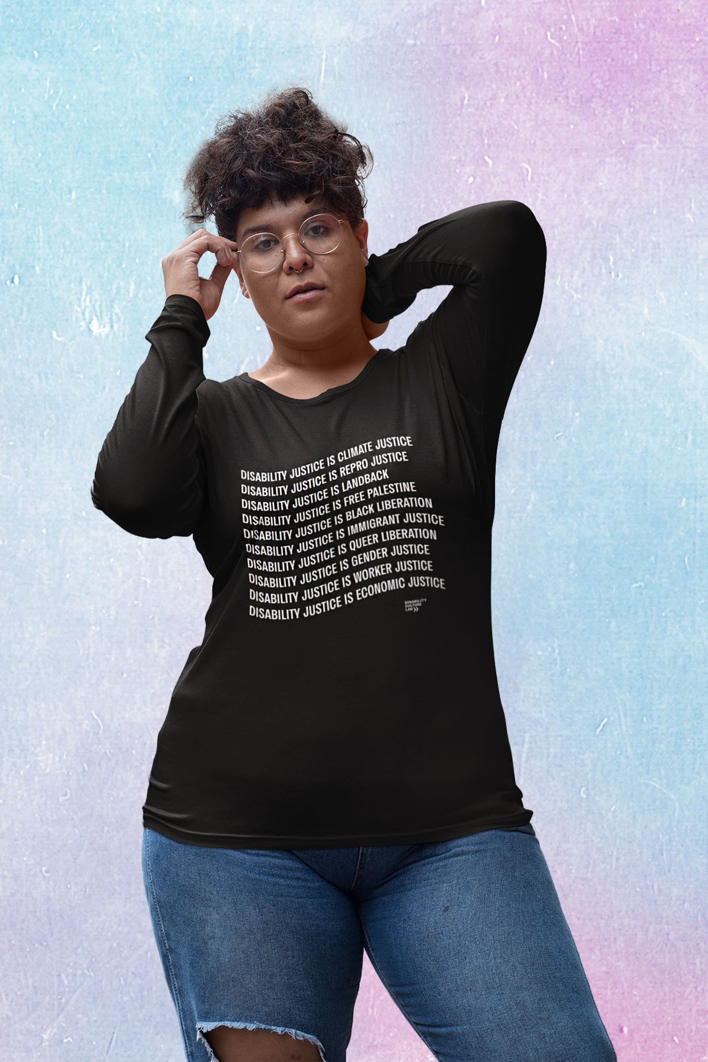 A person with curly brown hair poses holding their glasses and with a hand behind their head, lookking at the camera. They are wearing ripped blue jeans and a black long-sleeved shirt with white text that reads "Disability Justice Is... Climate Justice, Repro Justice, Landback, Free Palestine, Black Liberation, Immigrant Justice, Queer Liberation, Gender Justice, Worker Justice, Economic Justice" and the Disability Culture Lab logo in white. 