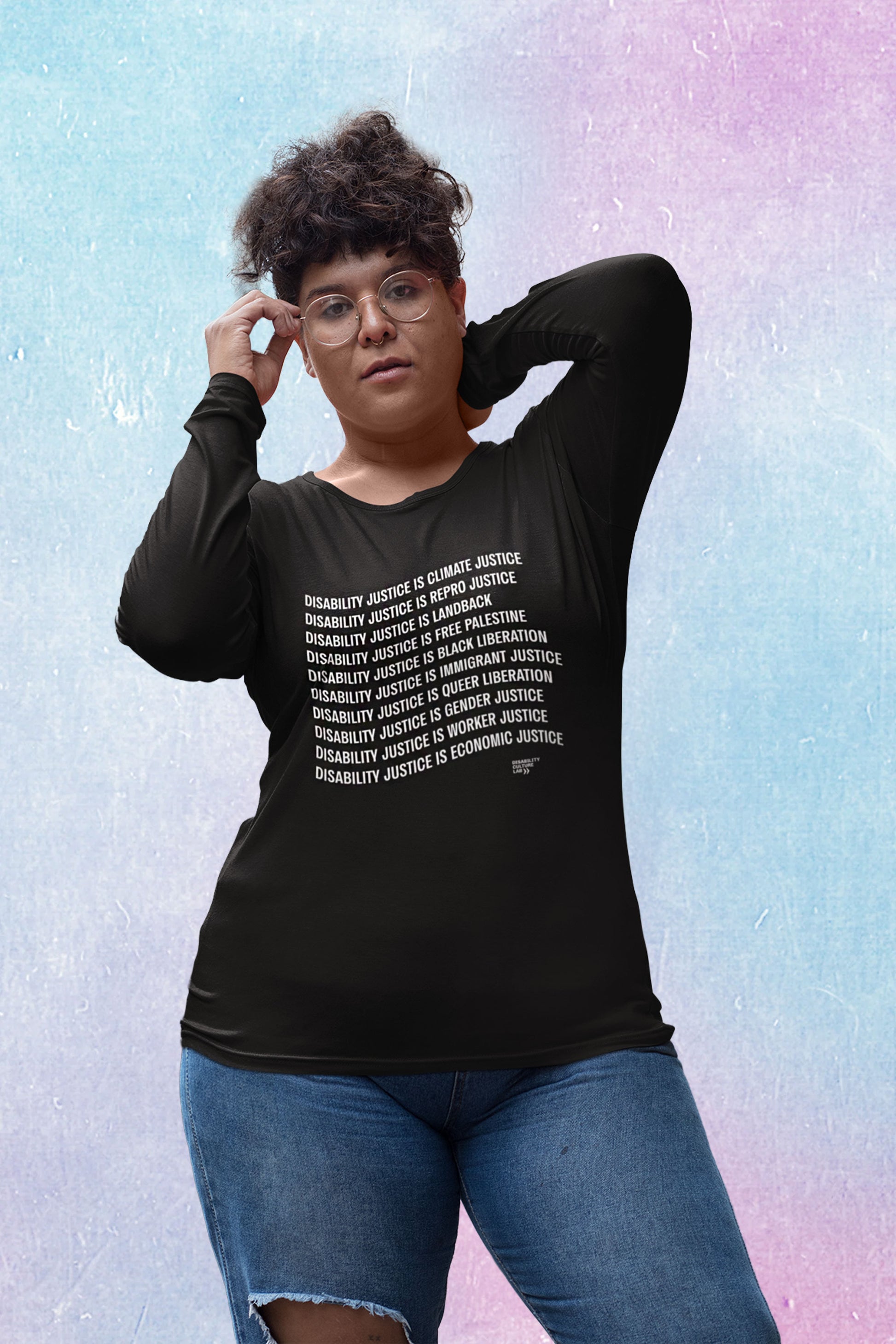 A person with curly brown hair poses holding their glasses and with a hand behind their head, lookking at the camera. They are wearing ripped blue jeans and a black long-sleeved shirt with white text that reads "Disability Justice Is... Climate Justice, Repro Justice, Landback, Free Palestine, Black Liberation, Immigrant Justice, Queer Liberation, Gender Justice, Worker Justice, Economic Justice" and the Disability Culture Lab logo in white. 