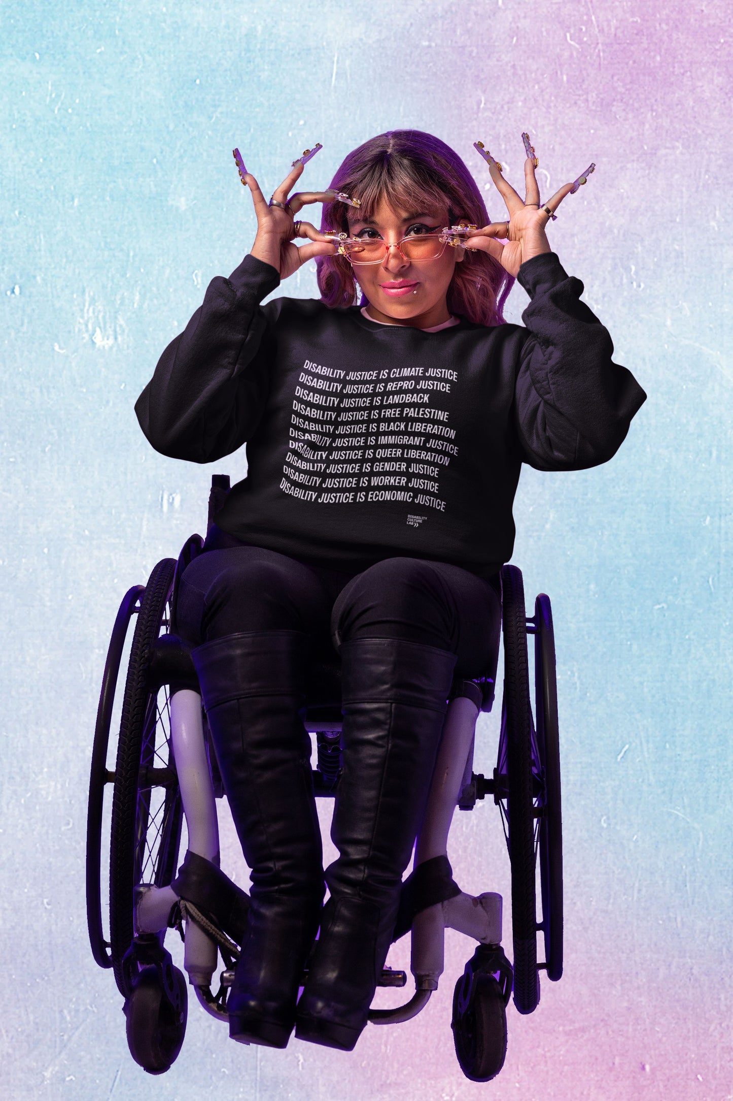 A brown-skinned person with brown hair and bangs, long nails, pink tinted sunglasses, and winged eyeliner lowers their glasses to look at the camera. They are seated in a wheelchair, wearing black knee-high boots, black pants, and a black crewneck sweater that reads "Disability Justice Is... Climate Justice, Repro Justice, Landback, Free Palestine, Black Liberation, Immigrant Justice, Queer Liberation, Gender Justice, Worker Justice, Economic Justice" and the Disability Culture Lab logo in white. They are s