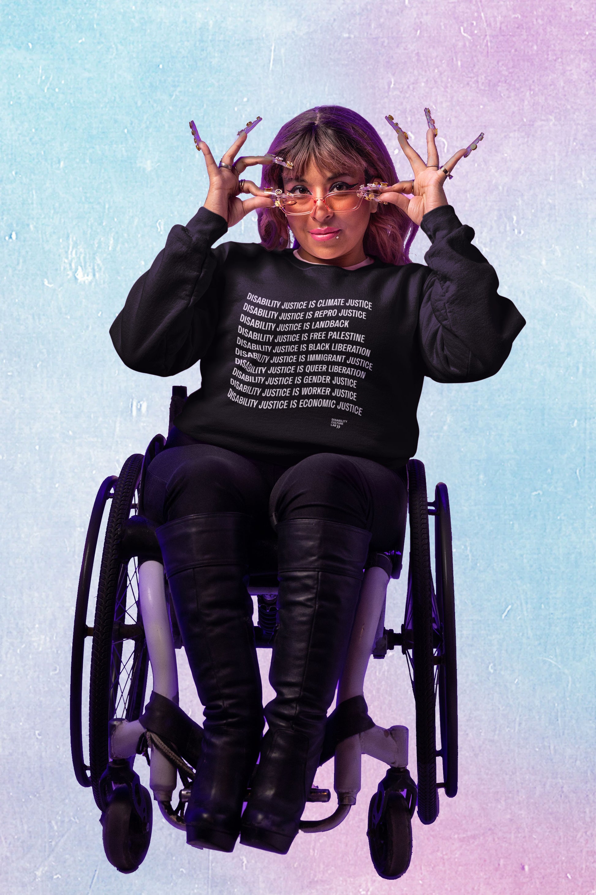 A brown-skinned person with brown hair and bangs, long nails, pink tinted sunglasses, and winged eyeliner lowers their glasses to look at the camera. They are seated in a wheelchair, wearing black knee-high boots, black pants, and a black crewneck sweater that reads "Disability Justice Is... Climate Justice, Repro Justice, Landback, Free Palestine, Black Liberation, Immigrant Justice, Queer Liberation, Gender Justice, Worker Justice, Economic Justice" and the Disability Culture Lab logo in white. They are s