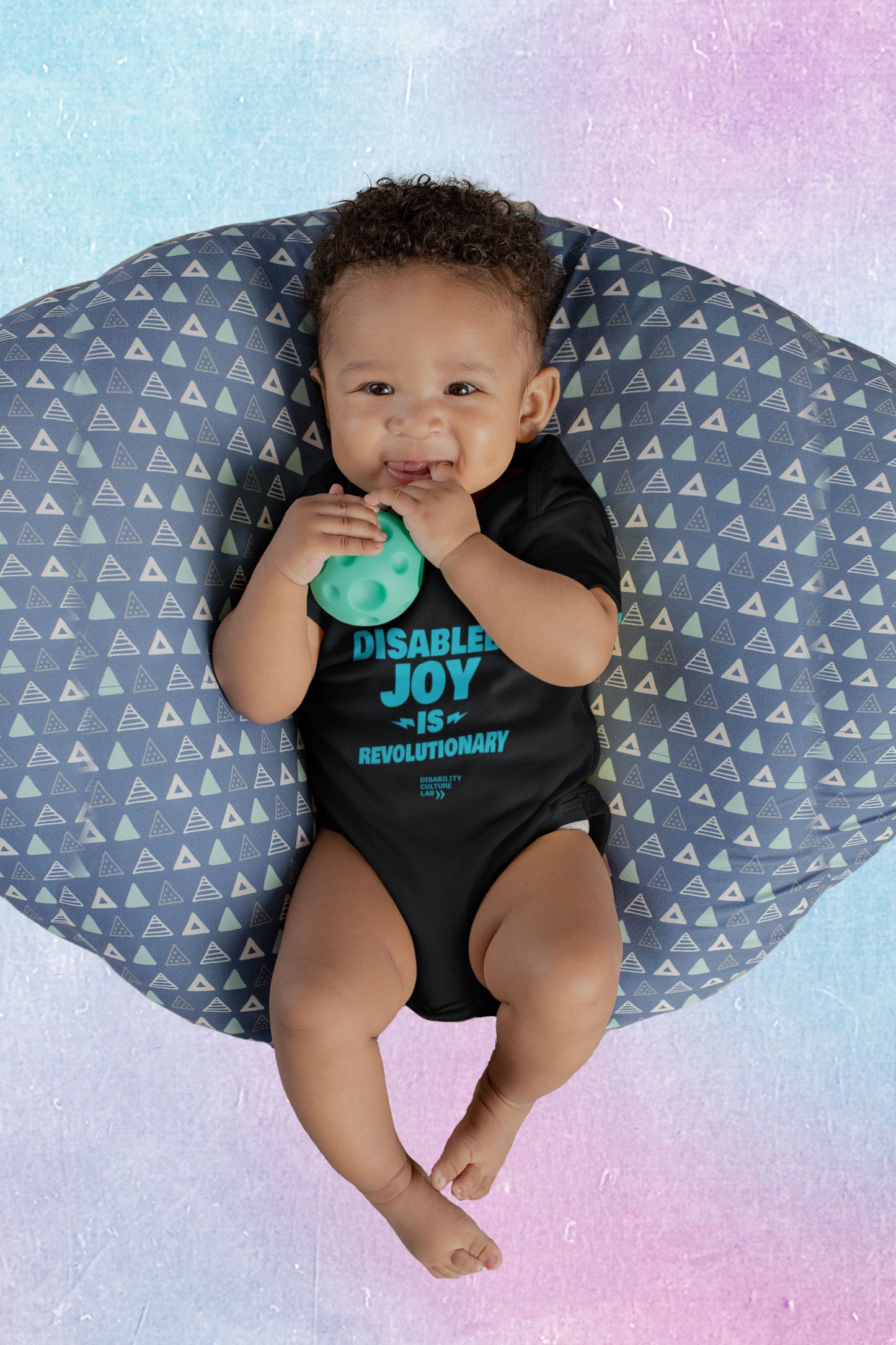 A very cute brown-skinned baby smiling and holding a green toy ball wearing a black onesie with the turquoise words "Disabled Joy Is Revolutionary" and the turquoise Disability Culture Lab logo. 