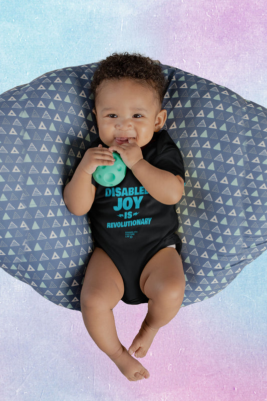 A very cute brown-skinned baby smiling and holding a green toy ball wearing a black onesie with the turquoise words "Disabled Joy Is Revolutionary" and the turquoise Disability Culture Lab logo. 