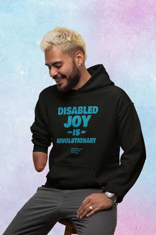 A person with blonde short-cropped hair and limb difference sits on a stool, looking down to the left and smiling. They are wearing gray pants, and a black pullover hoodie with the turquoise words "Disabled Joy Is Revolutionary" and the turquoise Disability Culture Lab logo.