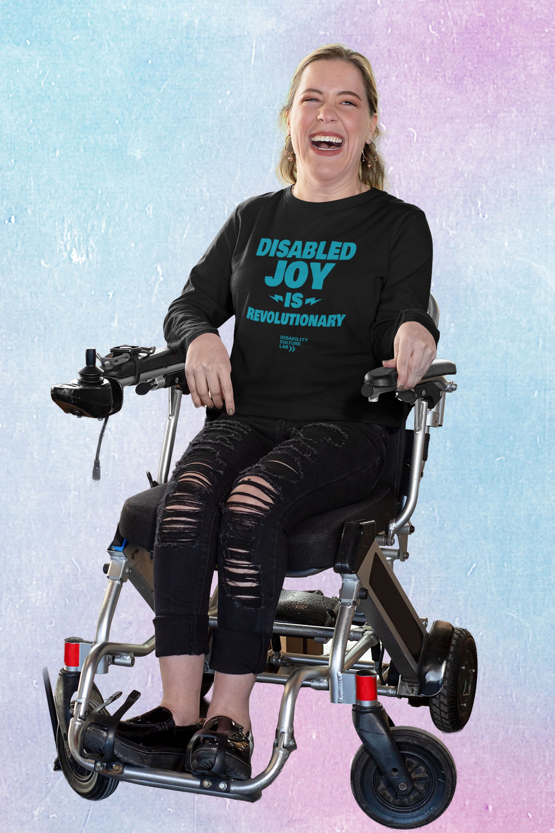 A person with brown hair sits in a power chair with a bright smile. They are wearing black ripped jeans, black shoes, and a black long-sleeved shirt with the turquoise words "Disabled Joy Is Revolutionary" and the turquoise Disability Culture Lab logo.