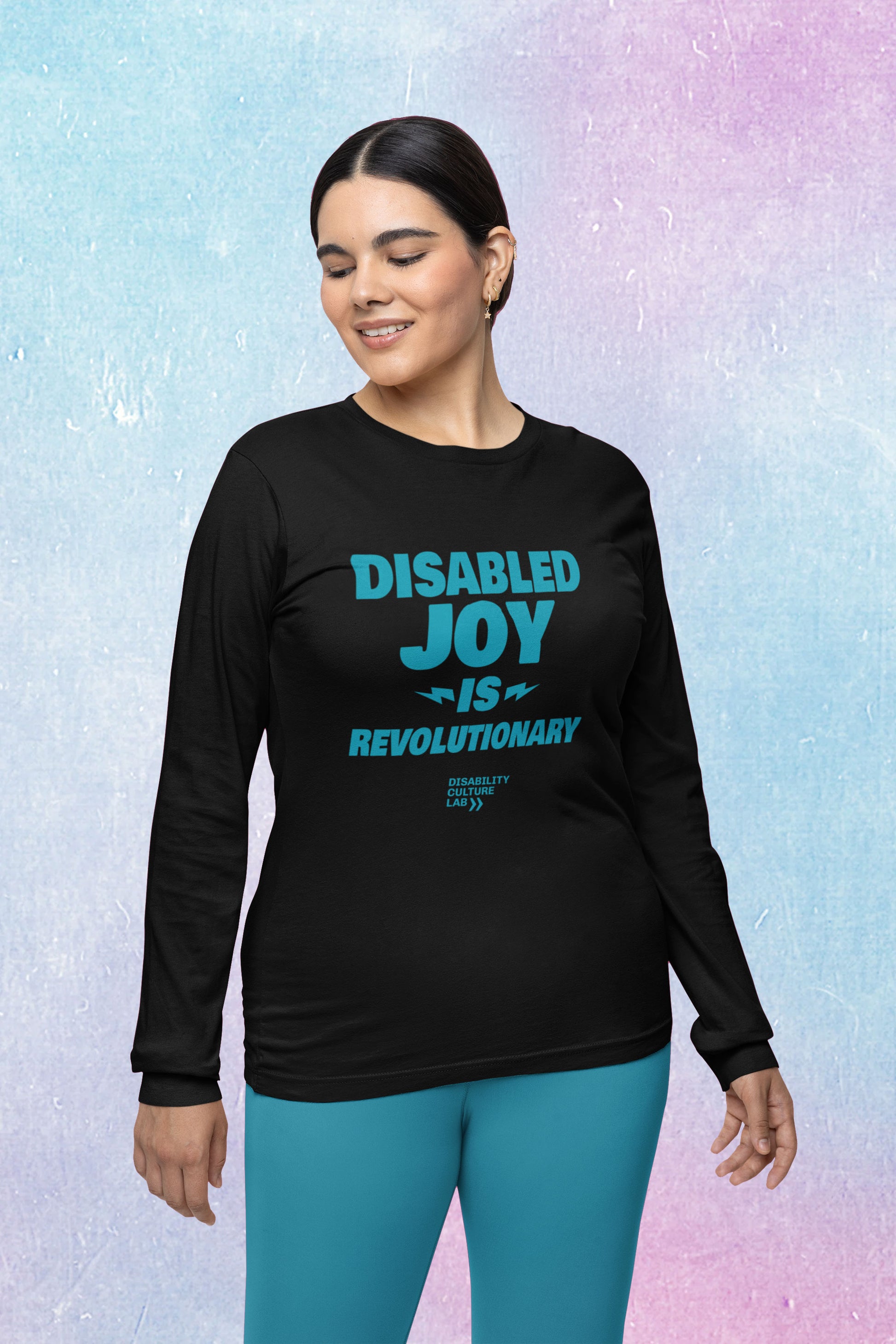 A light-skinned person with sleek black hair faces right with their eyes closed, wearing a turquoise leggings and a black long-sleeved shirt with the turquoise words "Disabled Joy Is Revolutionary" and the turquoise Disability Culture Lab logo. 