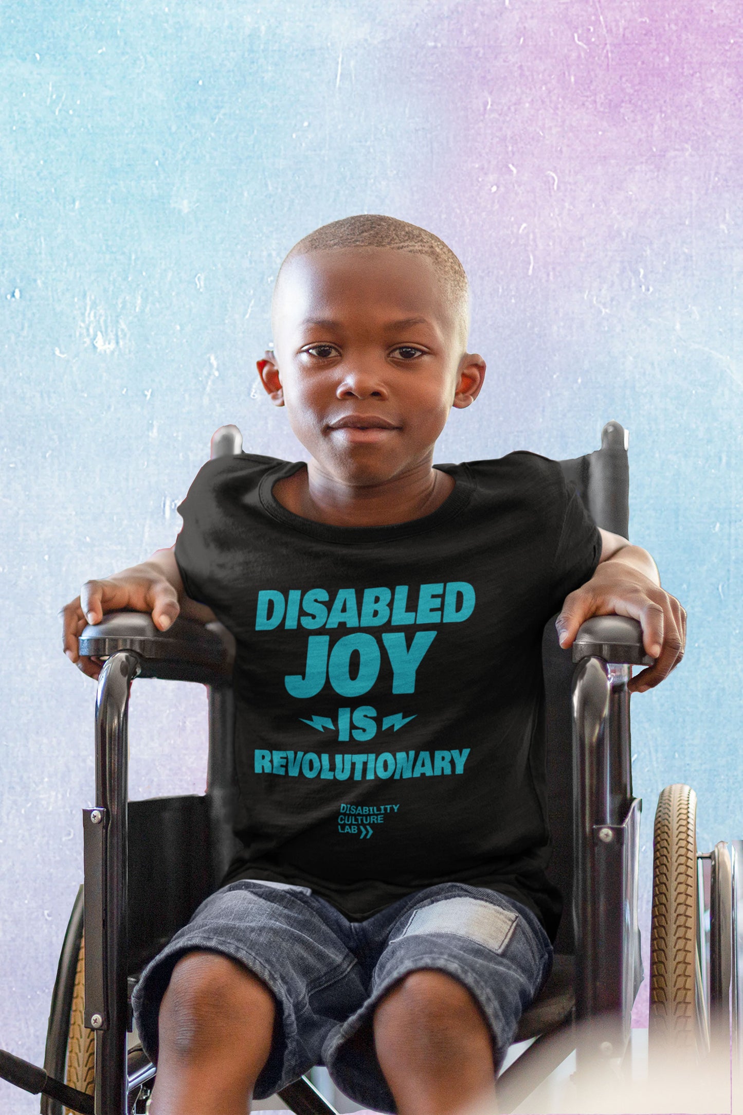 A very cute Black child sits in a wheelchair and looks at the camera, wearing blue jean shorts and a black t-shirt that reads "Disabled Joy Is Revolutionary" in turquoise, with the turquoise Disability Culture Lab logo below.