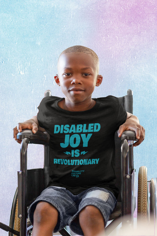 A very cute Black child sits in a wheelchair and looks at the camera, wearing blue jean shorts and a black t-shirt that reads "Disabled Joy Is Revolutionary" in turquoise, with the turquoise Disability Culture Lab logo below.