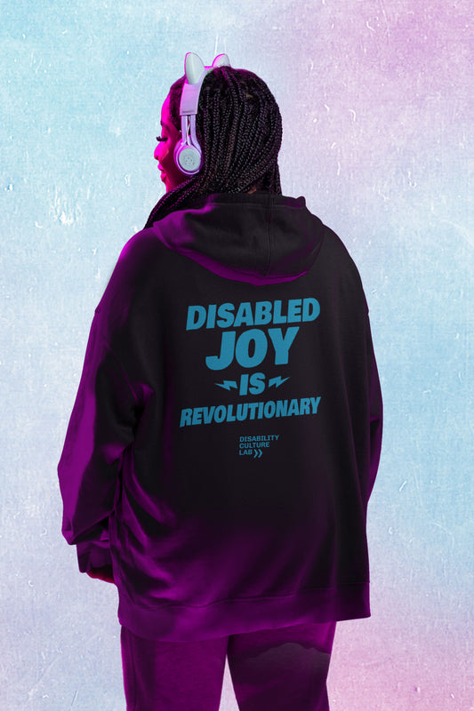 A Black person with long black braids and cute cat headphones faces away from the camera, showing the back of their black zip-up hoodie that reads "Disabled Joy Is Revolutionary" in turquoise, along with the turquoise Disability Culture Lab logo. They are illuminated in pink glowy light.