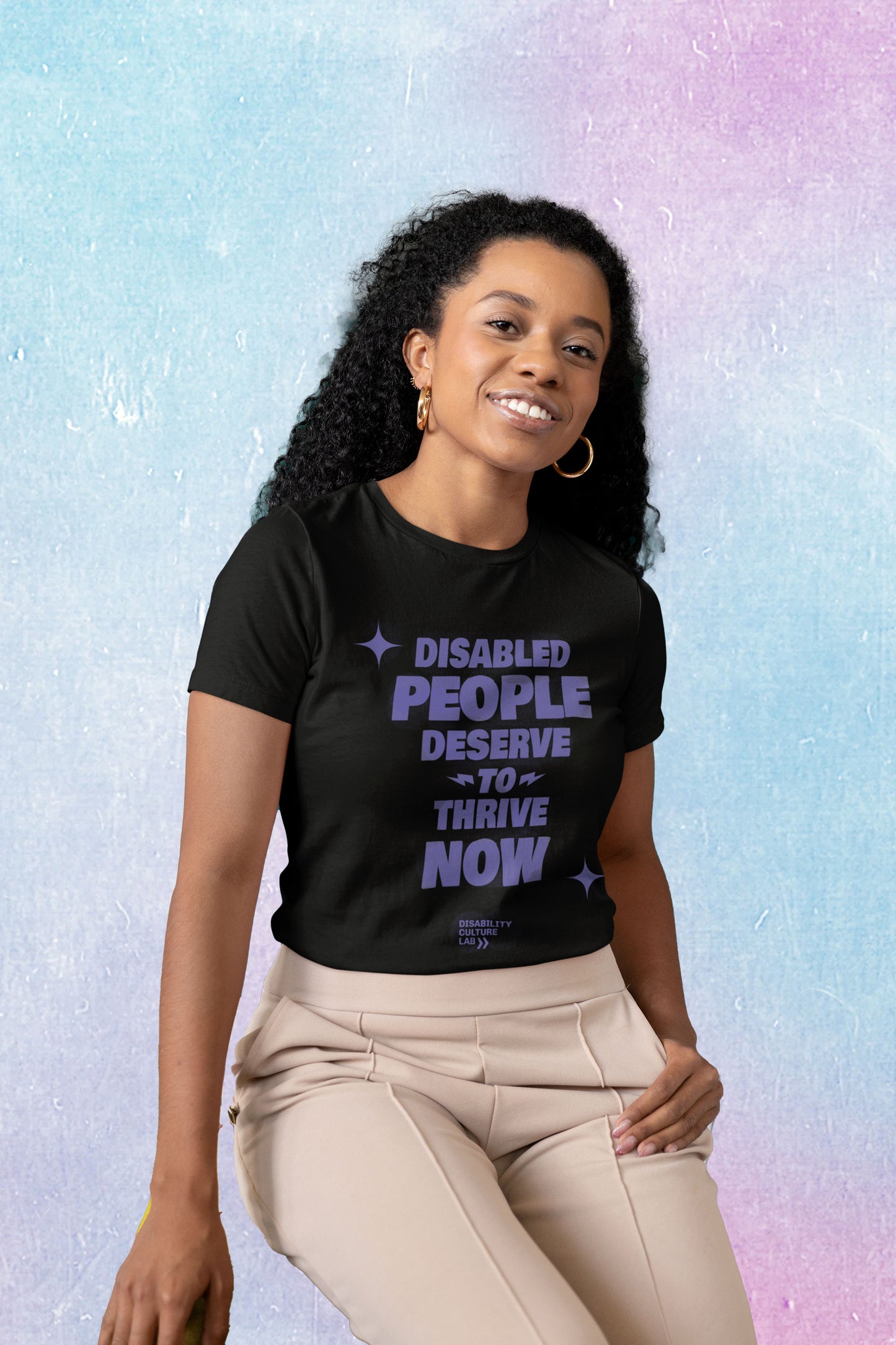 A Black person with long, black, curly hair, gold hoops, and tan pants tilts their head and smiles brightly at the camera. They are wearing a black t-shirt with the words "Disabled People Deserve To Thrive Now" in purple, with two purple sparkles, and the purple Disability Culture Lab logo below. 