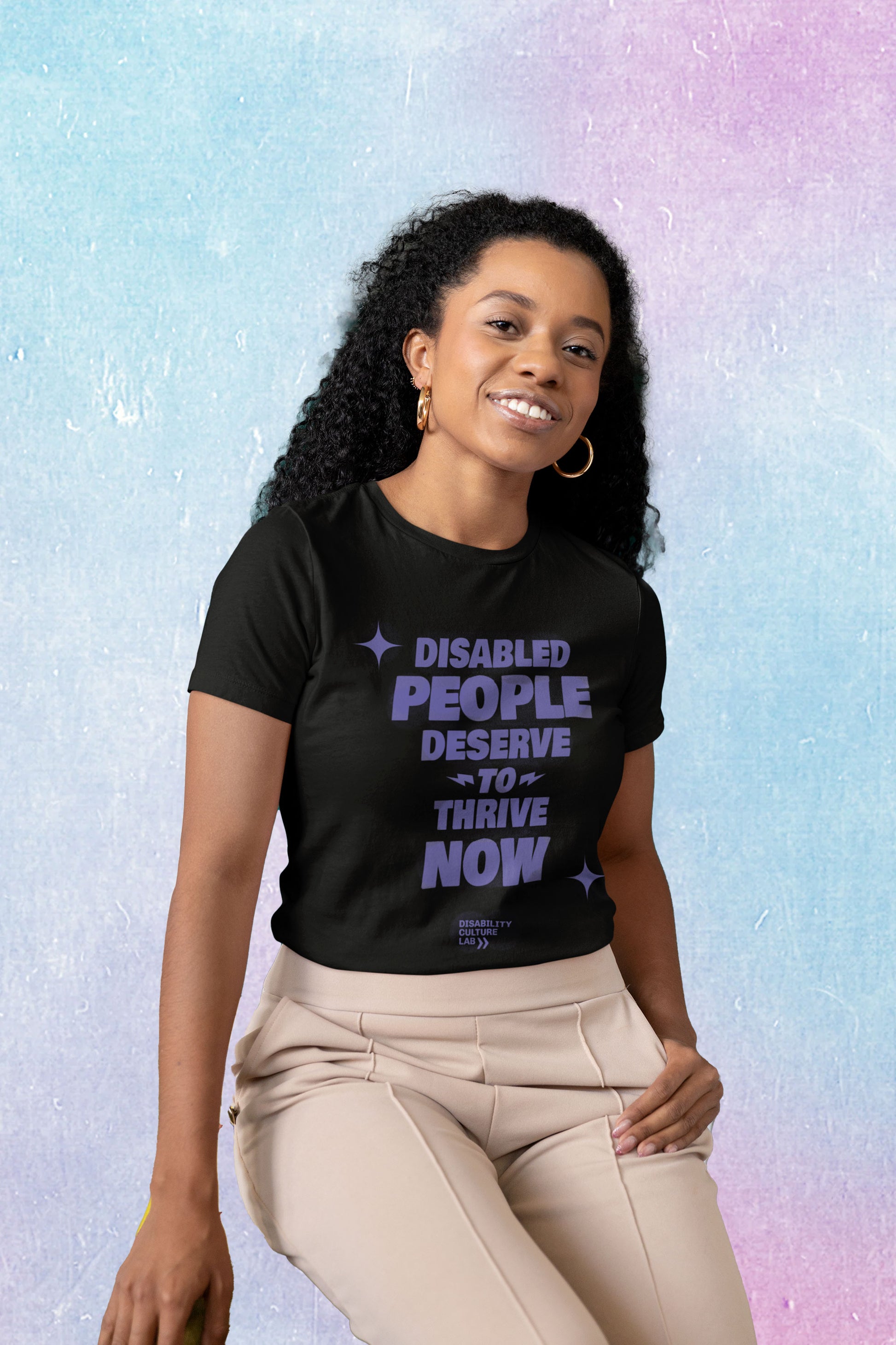 A Black person with long, black, curly hair, gold hoops, and tan pants tilts their head and smiles brightly at the camera. They are wearing a black t-shirt with the words "Disabled People Deserve To Thrive Now" in purple, with two purple sparkles, and the purple Disability Culture Lab logo below. 