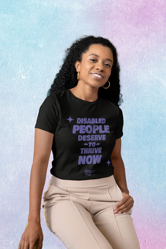 A Black person with long, black, curly hair, gold hoops, and tan pants tilts their head and smiles brightly at the camera. They are wearing a black t-shirt with the words "Disabled People Deserve To Thrive Now" in purple, with two purple sparkles, and the purple Disability Culture Lab logo below. 