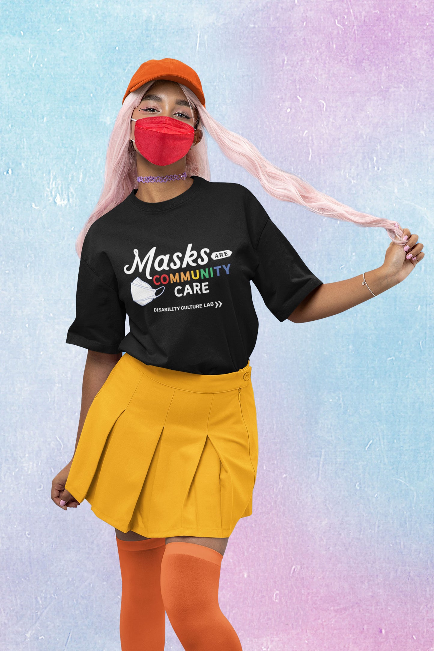 A person with long pale pink hair poses standing, looking at camera. They are wearing an orange hat, a red KF94 trifold respirator mask, a purple choker necklace, a yellow skirt, orange stockings, and a black t-shirt that reads "Masks Are Community Care," with "Masks" in a white handwritten font, "Are" in black text in a small white shape, "Community" in rainbow text, and "Care" in white text. To the left of the text is a white KF94 trifold respirator mask. Below is the white Disability Culture Lab logo.