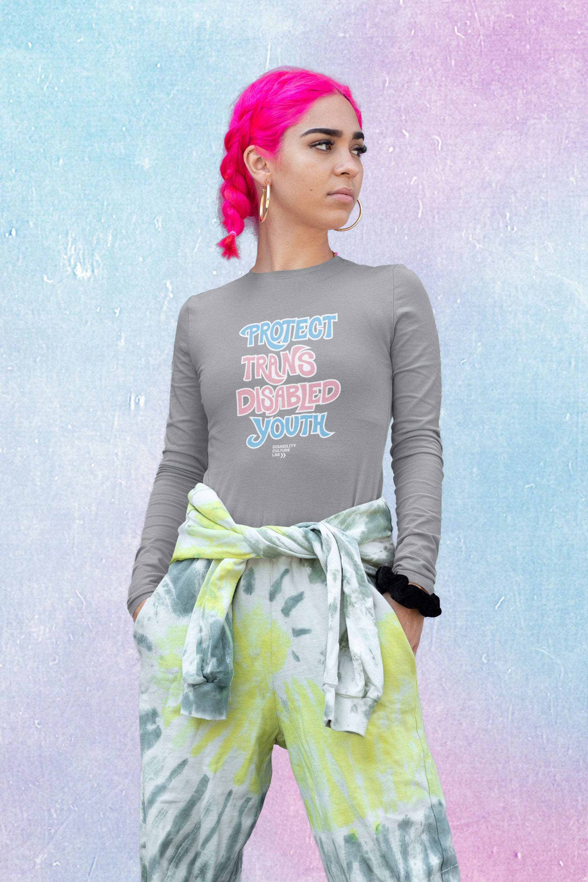 A light-skinned person with bright neon pink hair pulled back in two braids looks to the left. They are wearing gold hoop earrings. green and white tie-dye pants with a matching shirt tied around their waist, a black scrunchie on their wrist, and a long-sleeved gray shirt that reads “Protect Trans Disabled Youth” in hand drawn letters in the trans pride flag colors (light pink, sky blue, and white). Below is the white Disability Culture Lab logo.