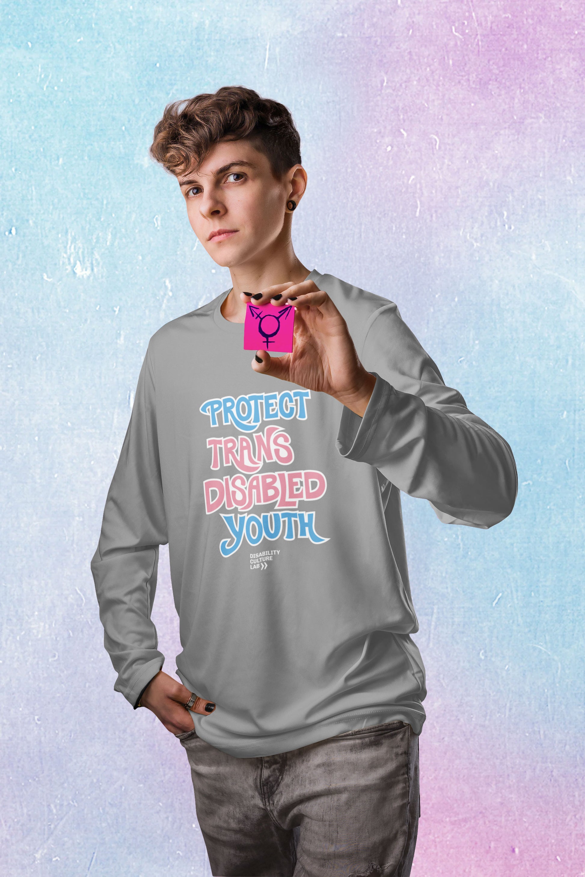 A white person with brown hair and shaved sides of their head looks into the camera while holding a small pink square with the black transgender symbol. They are wearing gray jeans and a long-sleeved  gray shirt that reads “Protect Trans Disabled Youth” in hand drawn letters in the trans pride flag colors (light pink, sky blue, and white). Below is the white Disability Culture Lab logo.