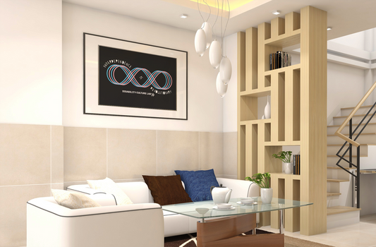 A framed Black "Interdependence is Revolutionary" poster on a wall. The room has white walls, a white couch with colorful pillows, a glass coffee table, lights hanging from the ceiling, and a wooden bookshelf. The poster has a design of a pink and blue four-line loop, like an infinity sign with an extra loop added to it. Text reads, "Interdependence Is Revolutionary" and starts at the top left of the diagram, weaves into the center of the diagram and then ends at the bottom right.