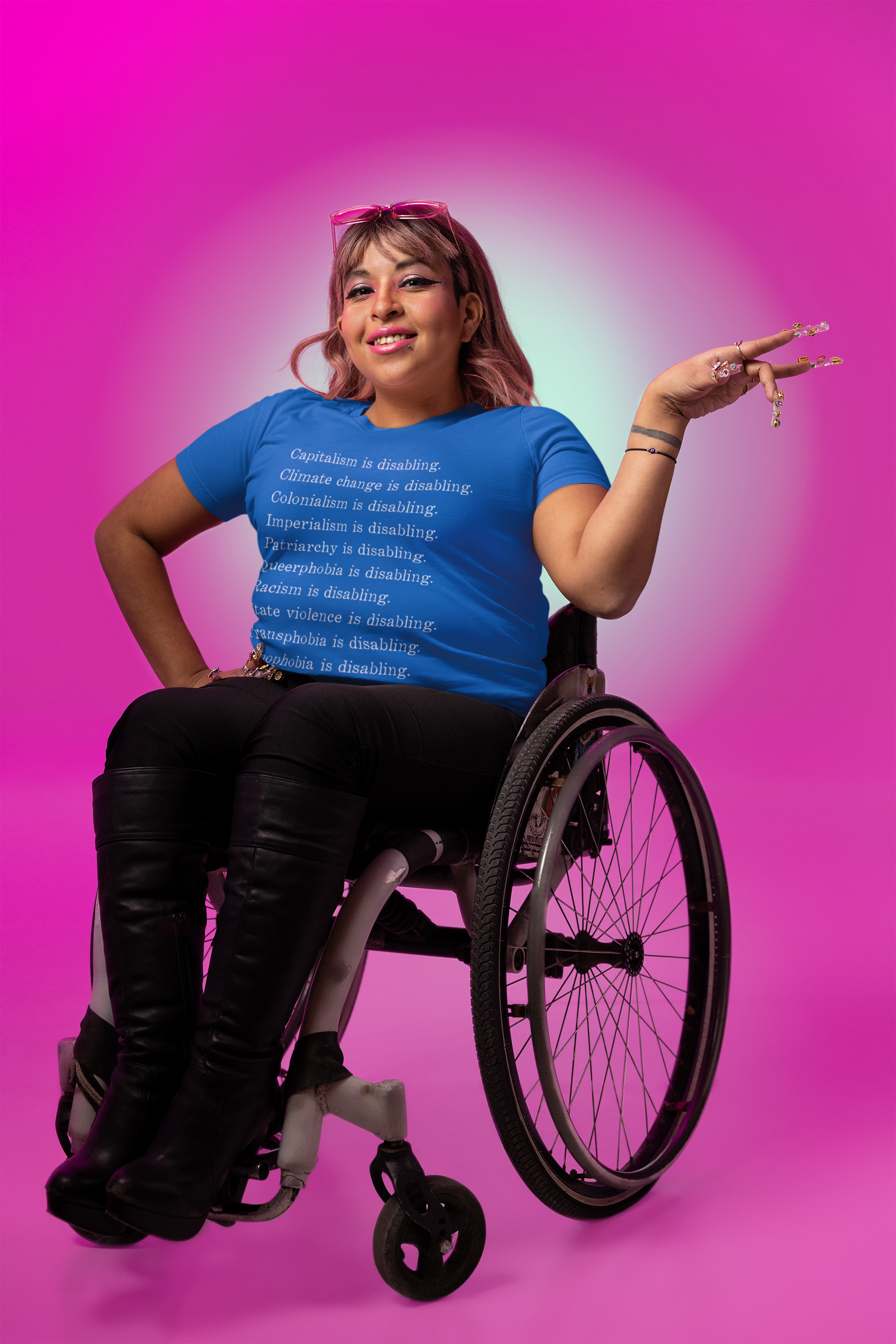 Light brown-skinned wheelchair user with sunglasses on their head wears a royal blue shirt that says "Oppression Is Disabling" t-shirt that says, "Racism is disabling. Capitalism is disabling. Colonialism is disabling. Transphobia is disabling. Climate change is disabling. State violence is disabling. Imperialism is disabling. Xenophobia is disabling. Queerphobia is disabling." 