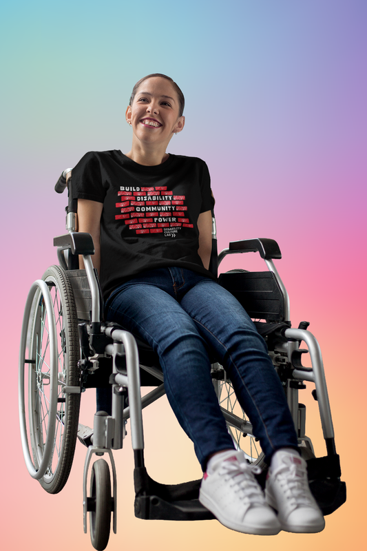 A white person sits in a wheelchair, wearing the "Build Disability Community Power" shirt, blue jeans, and white sneakers. The black short sleeved T-shirt has a design of red bricks in the middle of the shirt. The bricks are surrounding bold white text that reads "Build Disability Community Power." Beneath the bricks is the white Disability Culture Lab logo with two white arrows.
