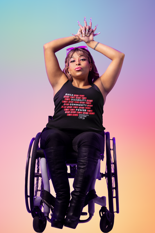 A person with light brown skin and shoulder-length hair sits in a wheelchair. They are wearing the black "Build Disability Community Power" muscle tank, with black pants and long black boots. Their arms are up above their head, and they have long glittery nails. The muscle tank has a design of red bricks in the middle of the shirt. The bricks are surrounding bold white text that reads "Build Disability Community Power." Beneath the bricks is the white Disability Culture Lab logo with two white arrows.