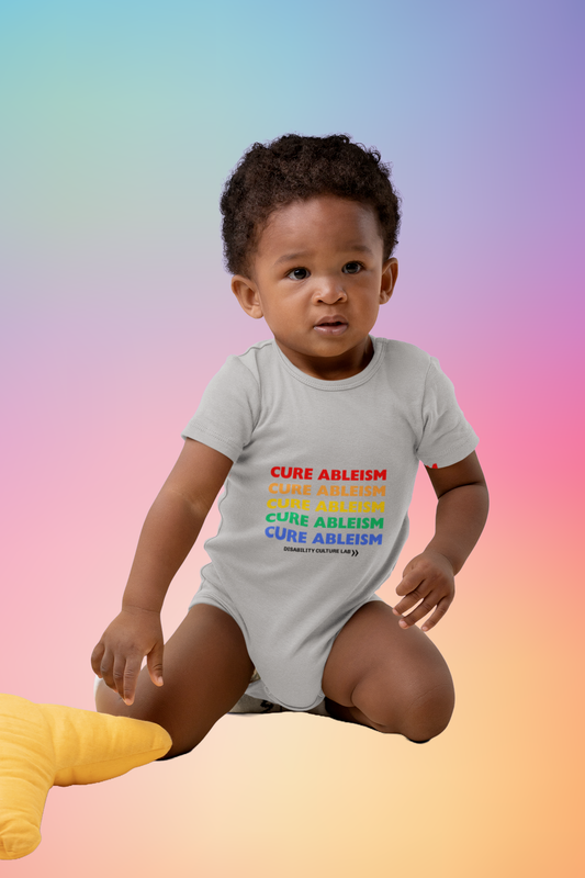 A very cute Black baby sits while wearing a gray onesie that reads "Cure Ableism" five times in red, orange, yellow, green, and blue, and the white Disability Culture Lab logo. 