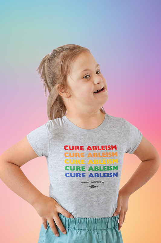 A very cute white child looks off to the side while wearing blue pants and a gray T-shirt that reads "Cure Ableism" five times in red, orange, yellow, green, and blue, and the white Disability Culture Lab logo. 