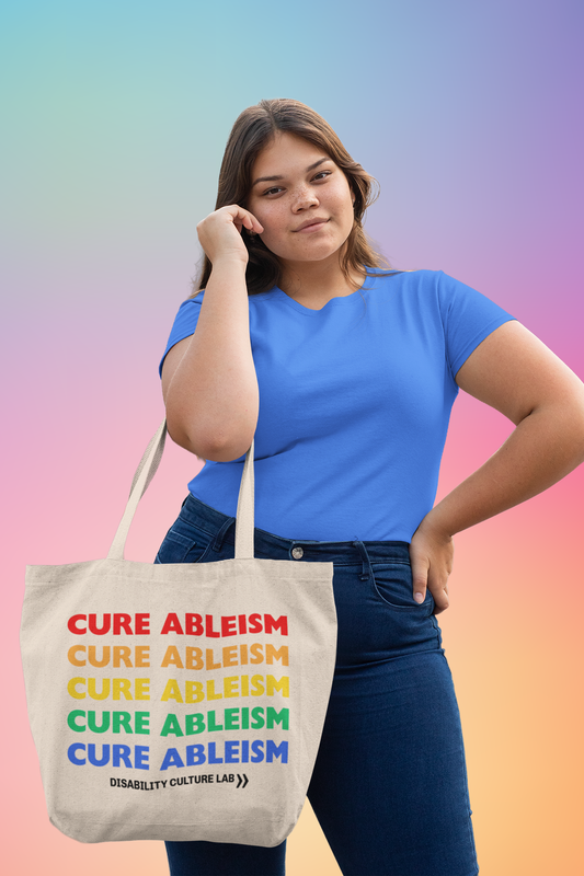 A light-skinned person with long brown hair wearing a blue shirt and blue jeans holding a beige tote bag that reads "Cure Ableism" five times in red, orange, yellow, green, and blue, and the white Disability Culture Lab logo. 