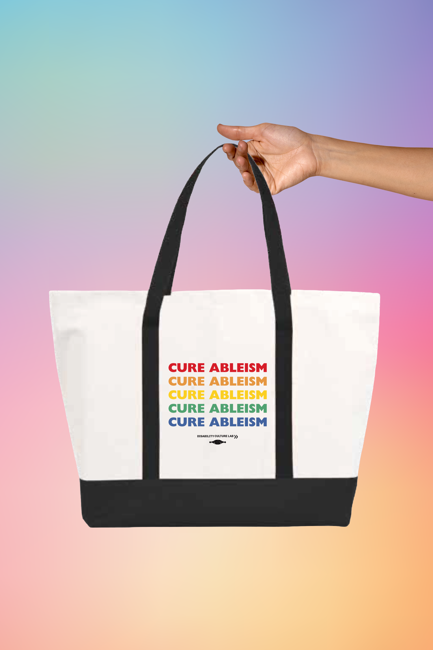 A hand holding A large white tote bag with black handles and bottom that reads "Cure Ableism" five times in red, orange, yellow, green, and blue, and the white Disability Culture Lab logo. 