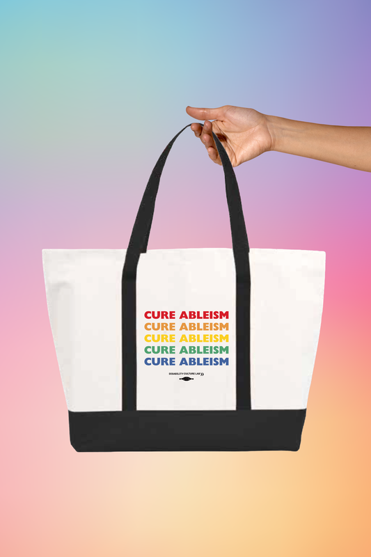 A hand holding A large white tote bag with black handles and bottom that reads "Cure Ableism" five times in red, orange, yellow, green, and blue, and the white Disability Culture Lab logo. 