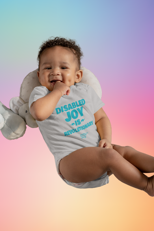 A very cute brown-skinned baby smiling sitting on an elephant pillow wearing a gray onesie with the turquoise words "Disabled Joy Is Revolutionary" and the turquoise Disability Culture Lab logo. 