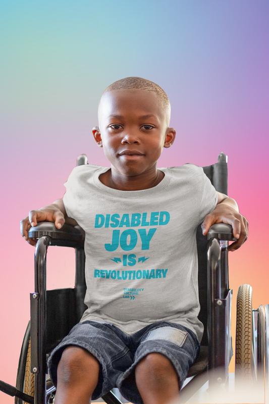 A very cute Black child sits in a wheelchair and looks at the camera, wearing blue jean shorts and a gray t-shirt that reads "Disabled Joy Is Revolutionary" in turquoise, with the turquoise Disability Culture Lab logo below.